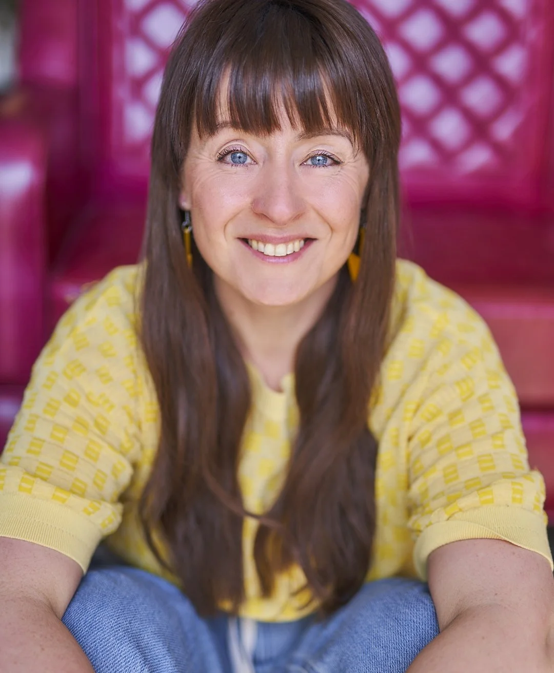 Leesa is leaning over her knees, sitting on the floor, smiling at the camera. She has long brown hair and is wearing a yellow t-shirt and blue jeans.