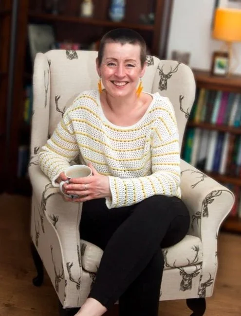 Leesa is sitting in a wingback chair, holding a mug of tea, smiling at the camera, face on. There are book shelves full of books in the background.