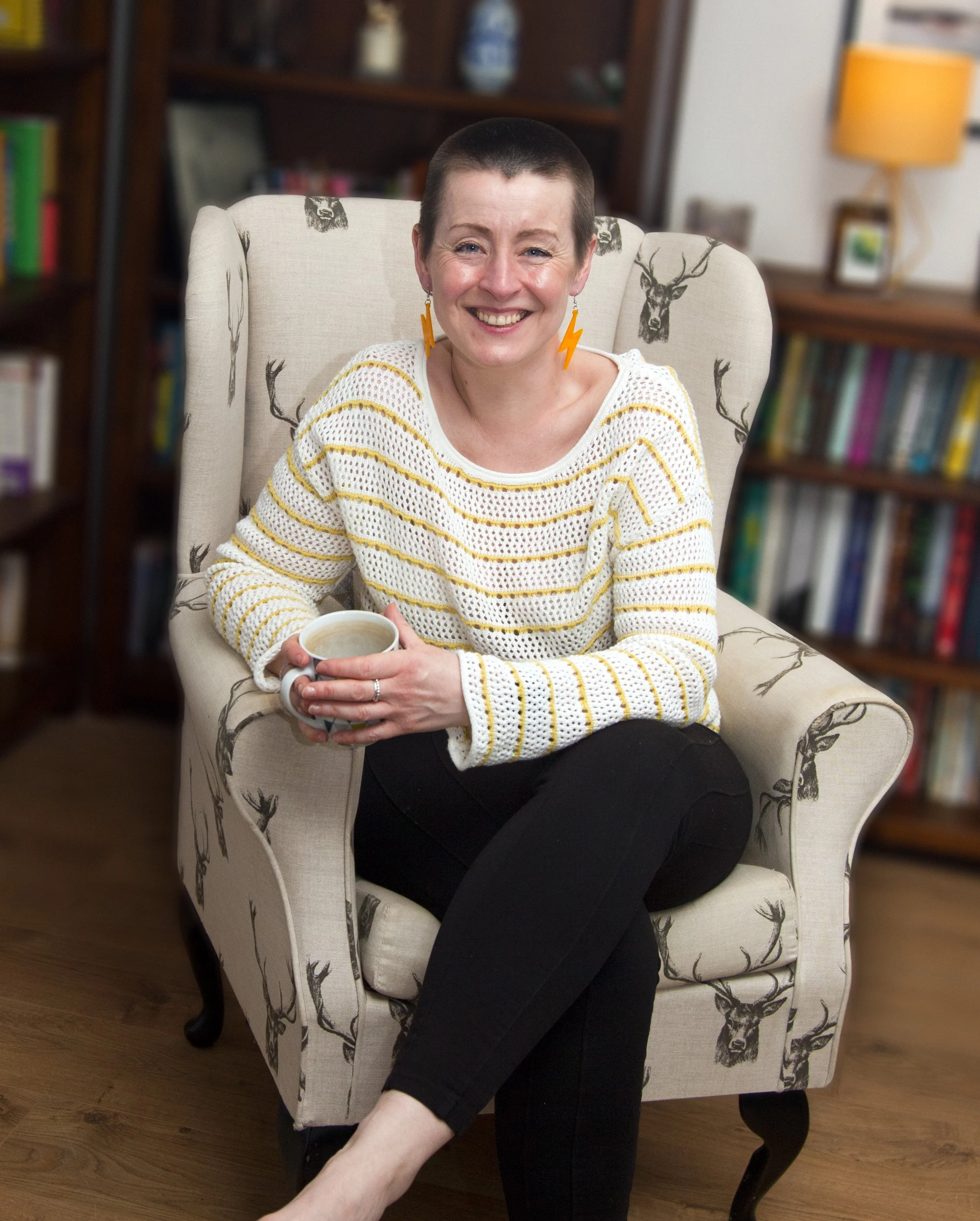 A smiling woman with short hair wearing a white and yellow striped sweater, sitting in a patterned armchair, holding a coffee mug, in a room with bookshelves and a lamp in the background.