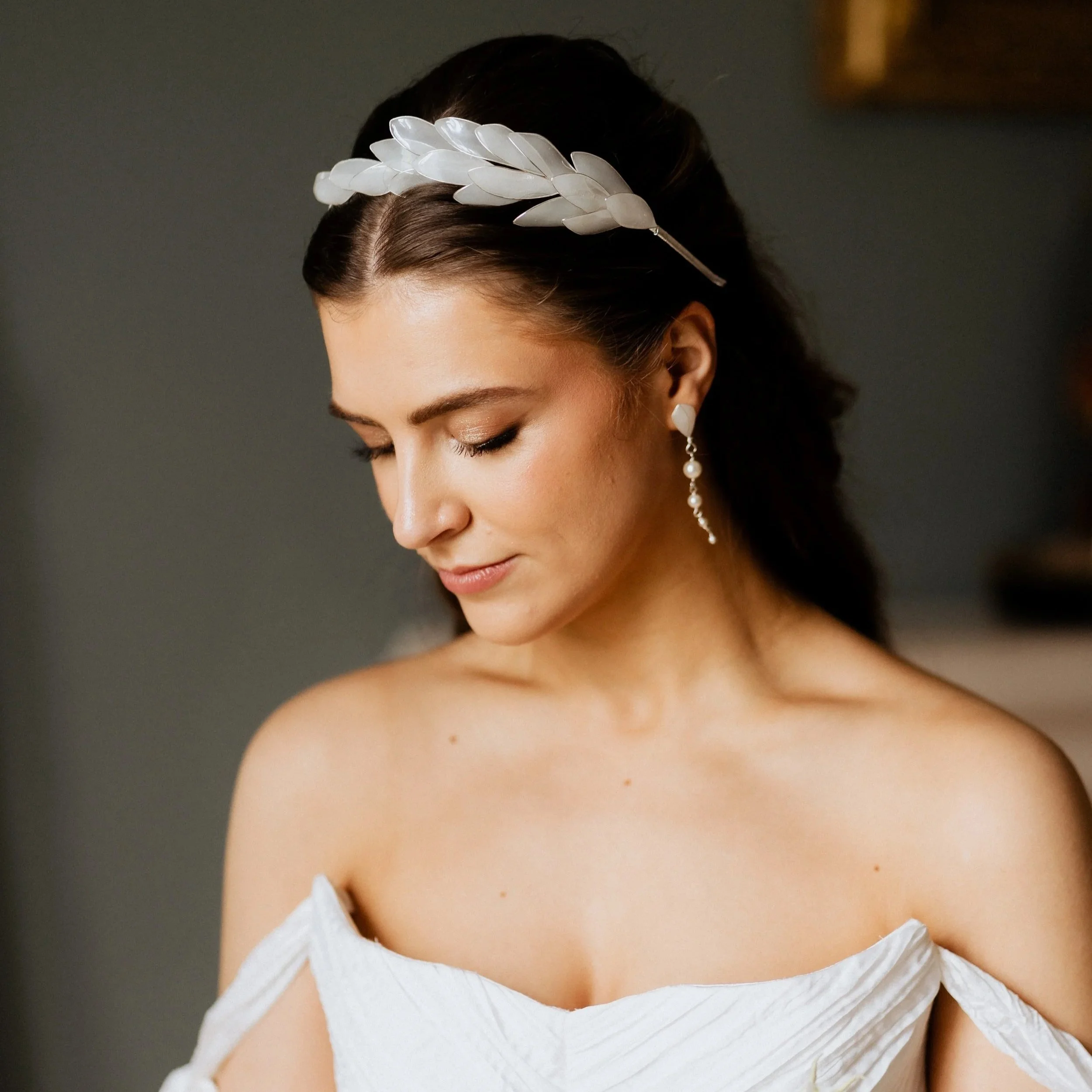 A woman with dark hair styled back, wearing a white off-the-shoulder dress, a white leaf couture headband by Jen Gilbert Bridal Accessories, and long dangling pearl earrings, looking downward with a soft expression.