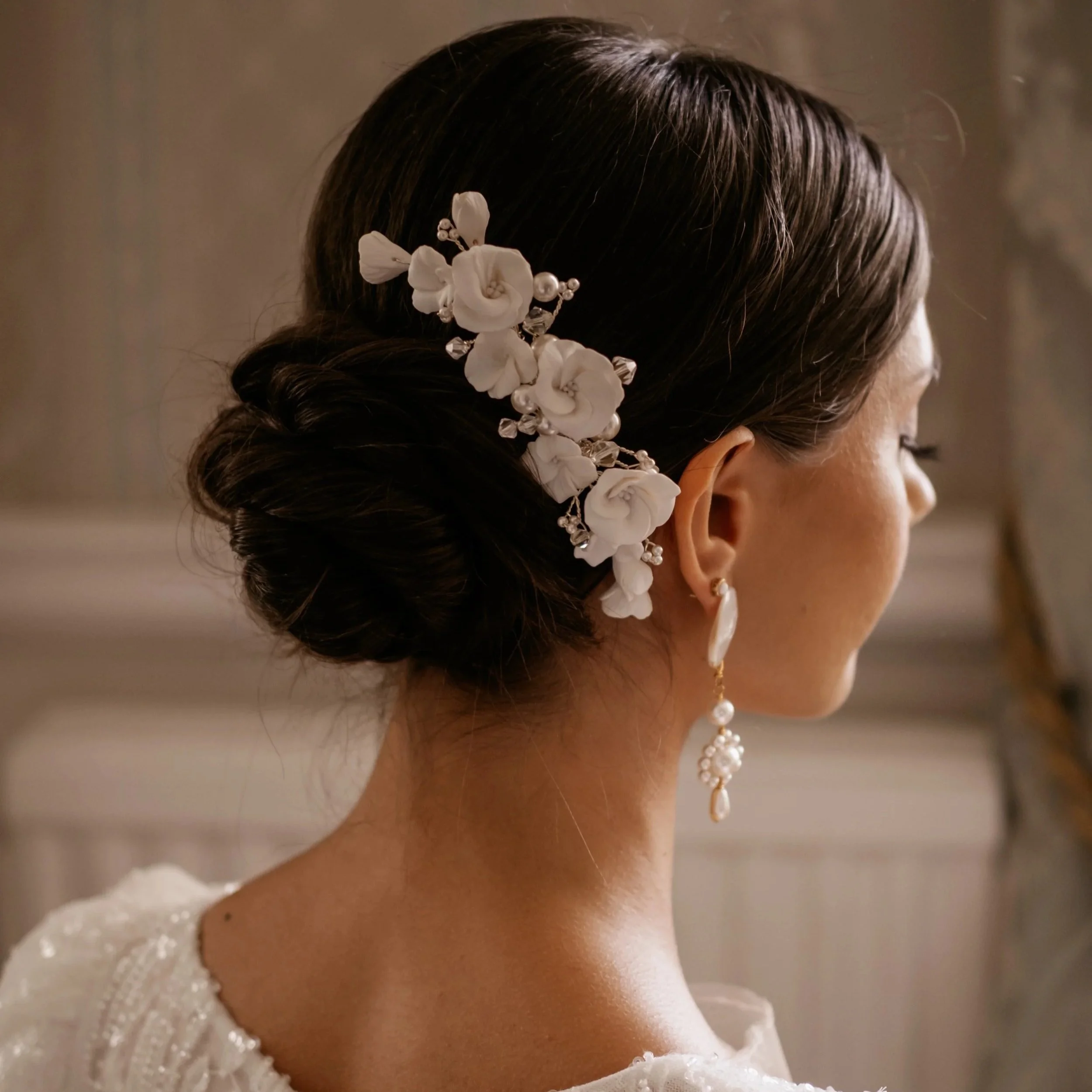 A woman with dark hair styled in an elegant updo, adorned with a white floral hairpiece and pearl accents by Jen Gilbert Bridal Accessories, wearing large pearl earrings and a white dress.