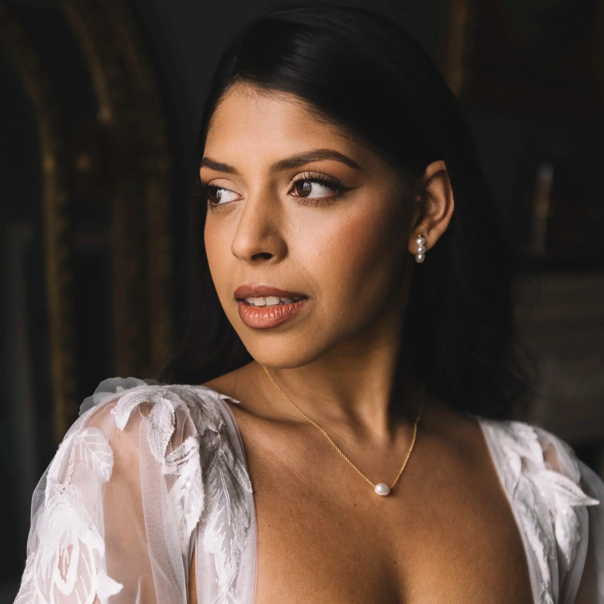 Close-up of a woman with dark hair, wearing pearl earrings and a pearl necklace by Jen Gilbert Bridal Accessories, dressed in a sheer white lace gown with floral embroidery.