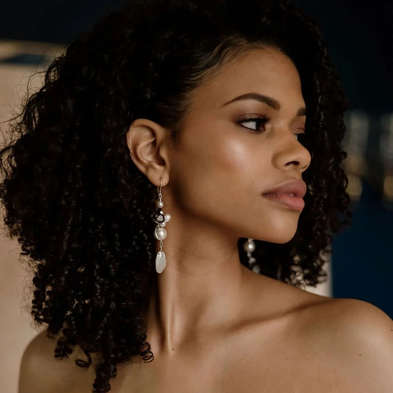 Close-up of a woman with curly dark hair wearing pearl and mother of pearl drop earrings by Jen Gilbert Bridal Accessories, looking to the side.