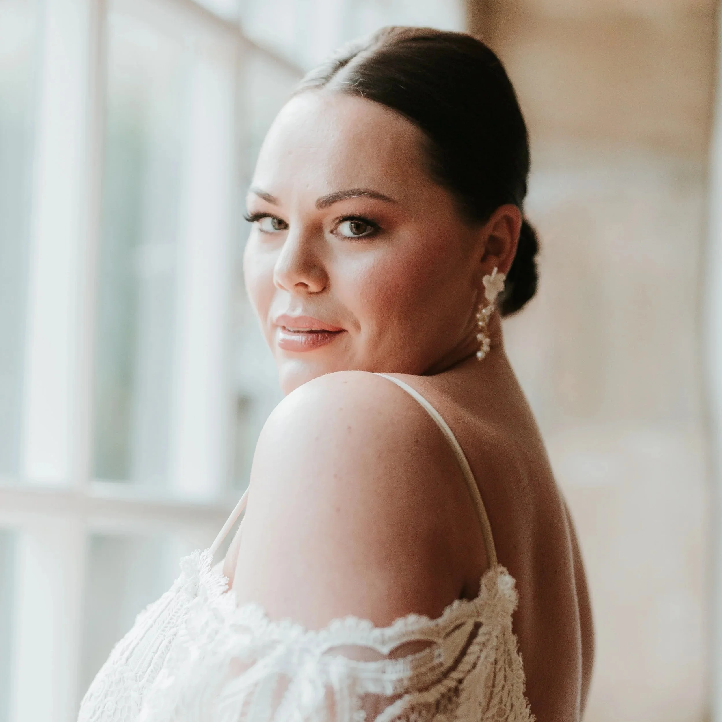 A woman with dark hair styled in an elegant bun, wearing a white lace dress with thin straps and statement earrings by Jen Gilbert Bridal Accessories, looking over her shoulder near a window with natural light.