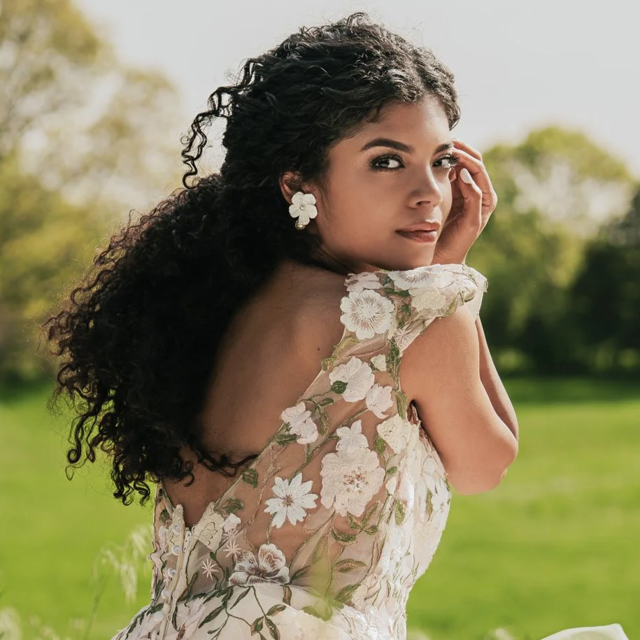 A woman with curly dark hair and floral earrings poses outdoors in a floral dress with lace details, surrounded by greenery and trees. Wearing statement earring by Jen Gilbert Bridal Accessories