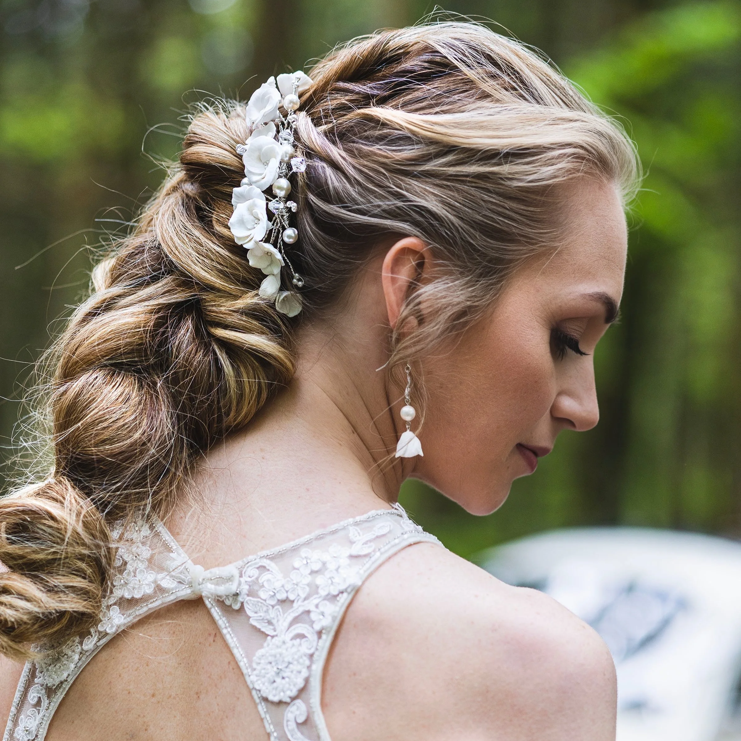 A bride with styled hair adorned with white flowers and pearls, wearing drop earrings and a lace wedding dress, is captured outdoors with a blurred green forest background, her eyes closed and head slightly tilted.