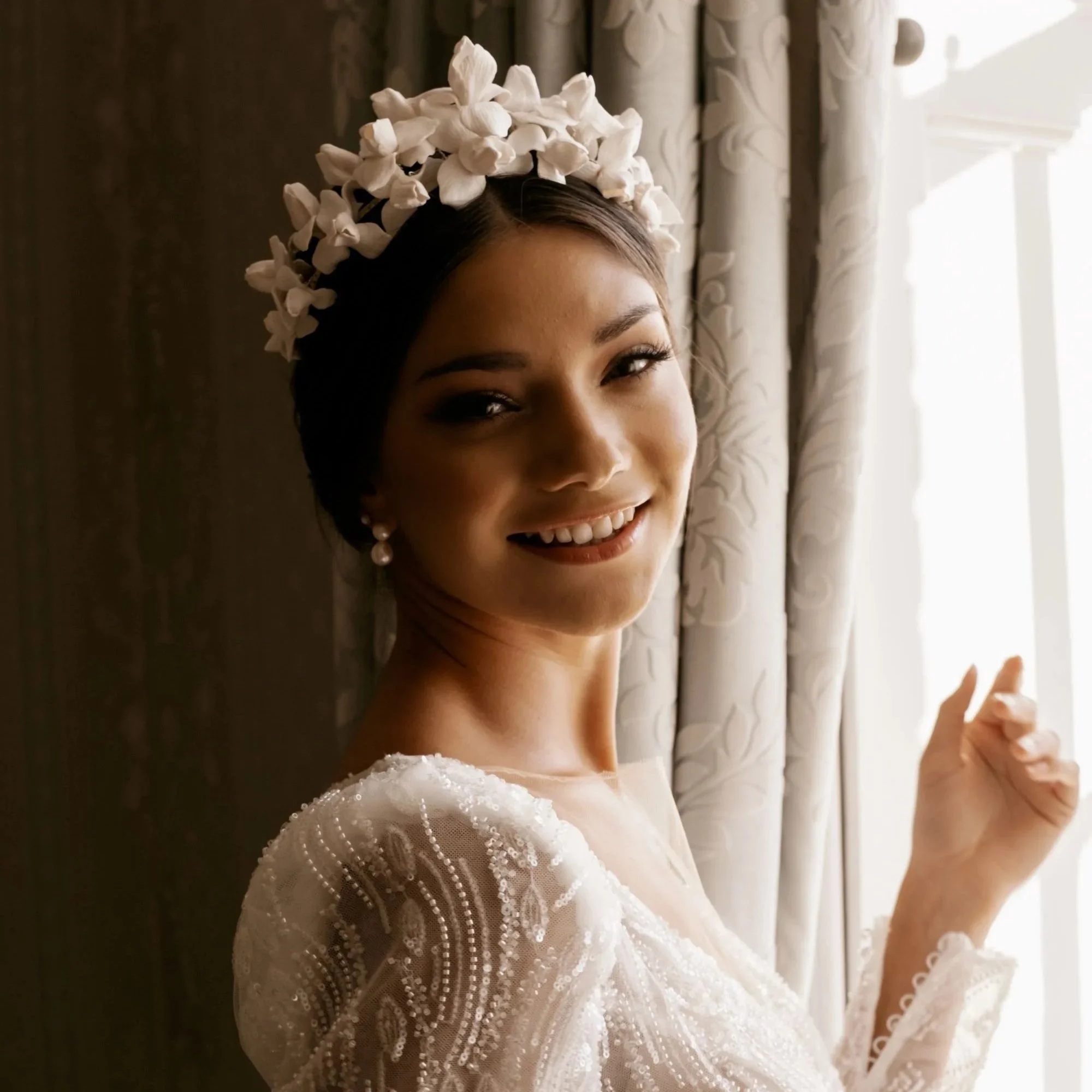 A bride with dark hair, wearing a white dress with beaded details, smiling and standing by a window with floral curtains, wearing a floral headpiece and pearl earrings by Jen Gilbert Bridal Accessories.