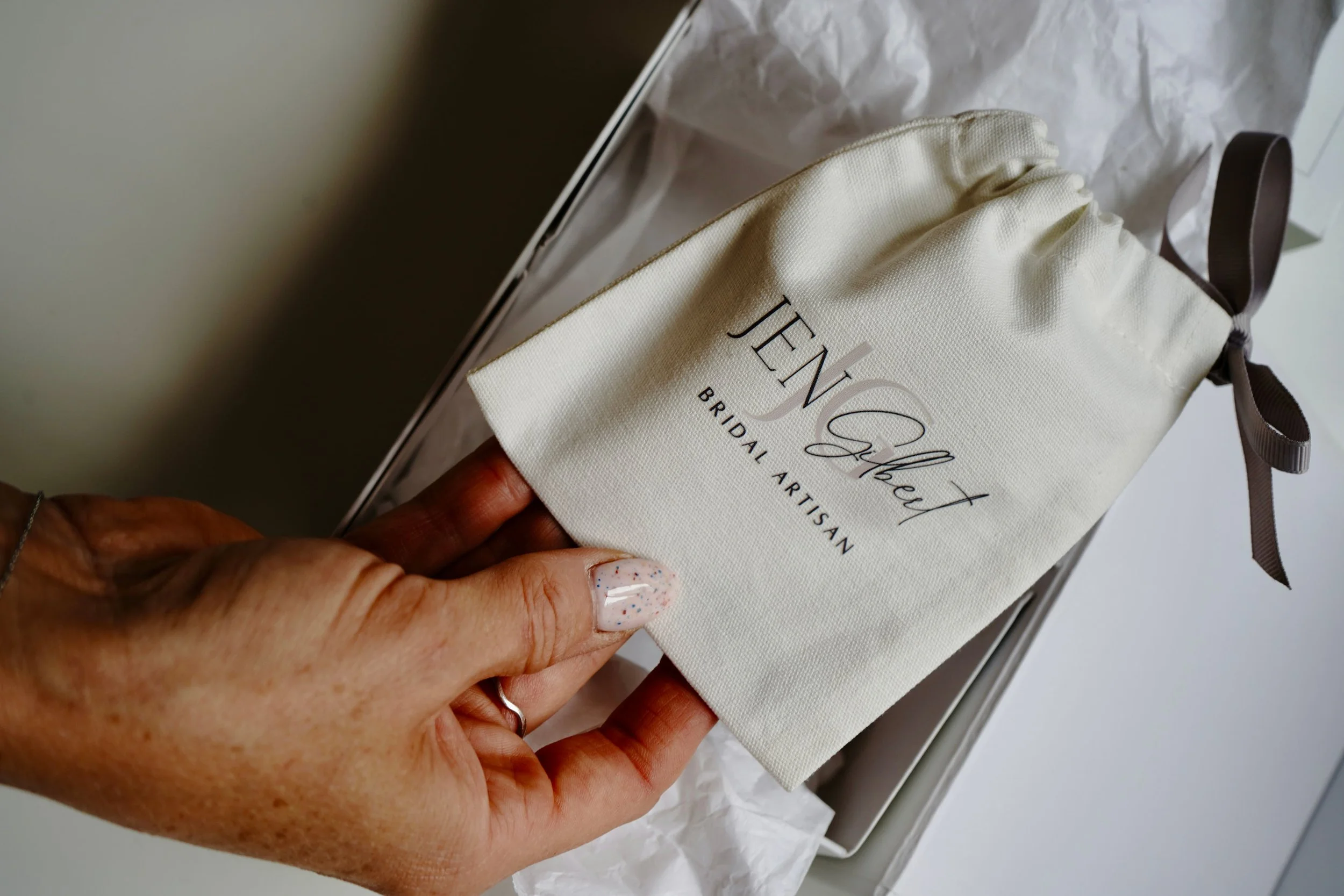 Hand holding a branded linen drawstring pouch by Jen Gilbert Bridal Artisan, presented in luxury packaging with tissue paper—elegant bridal accessory packaging detail.