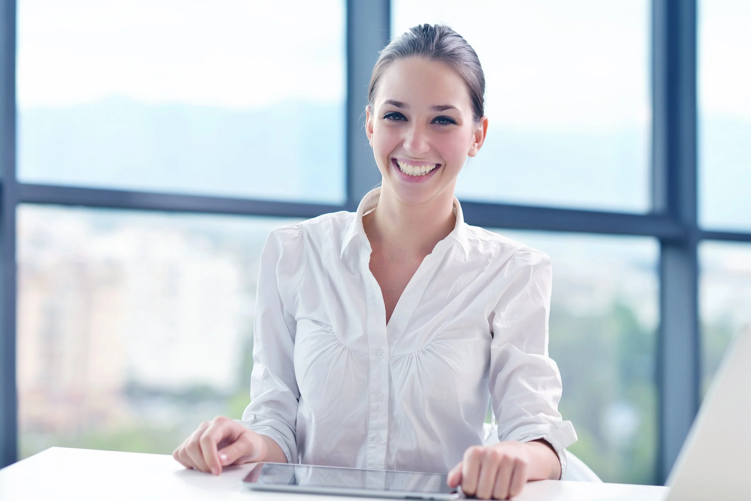 A young woman with brown hair sitting at a desk in front of large windows, smiling at the camera, with a tablet and a laptop in front of her.