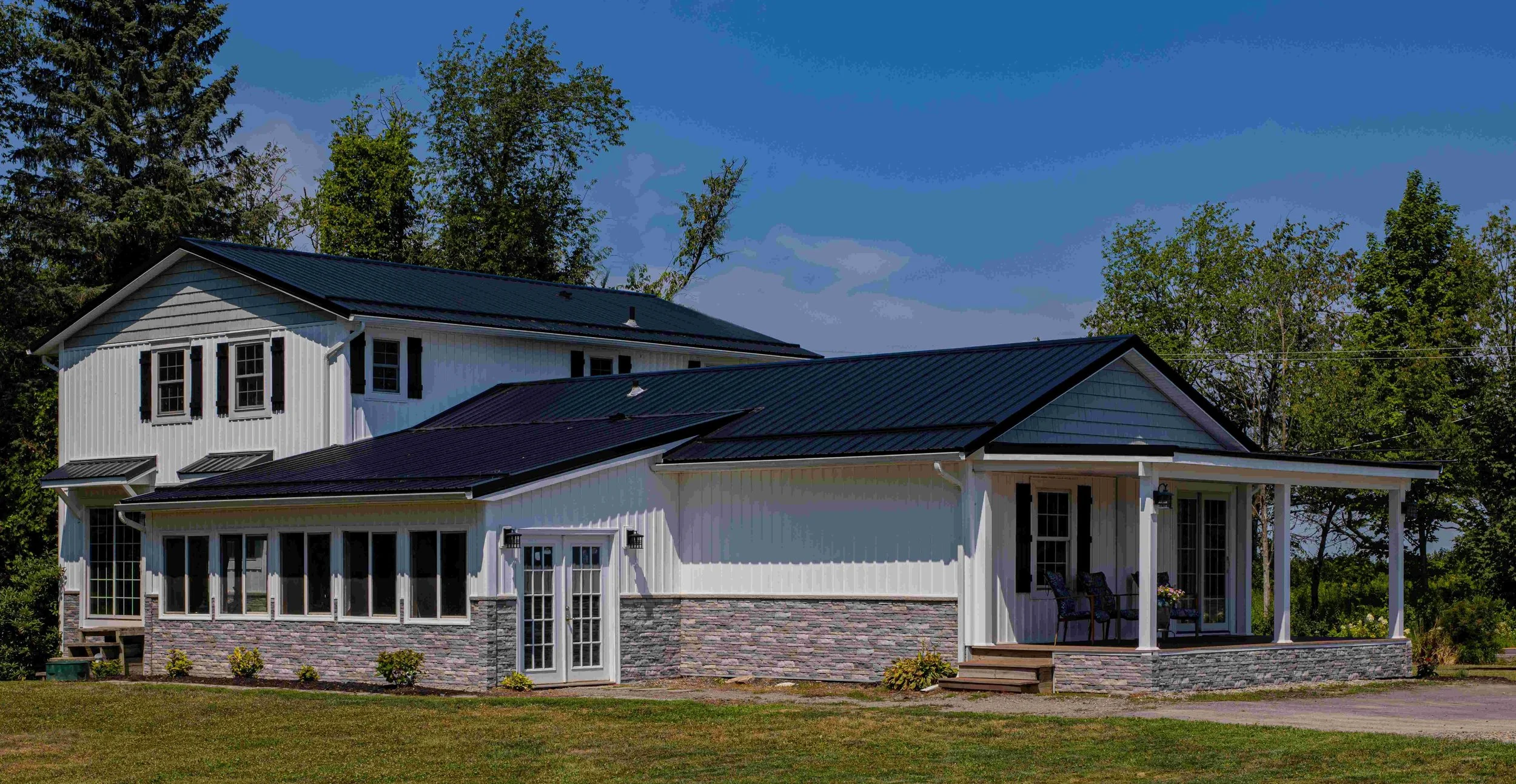 A two-story house with white siding, black metal roof, a screened porch with patio furniture, and a green lawn, surrounded by trees under a clear blue sky.