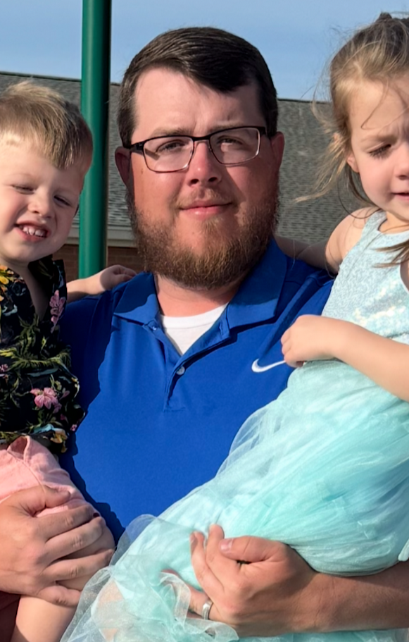 A man with glasses and a beard holding two young children outdoors during daytime.