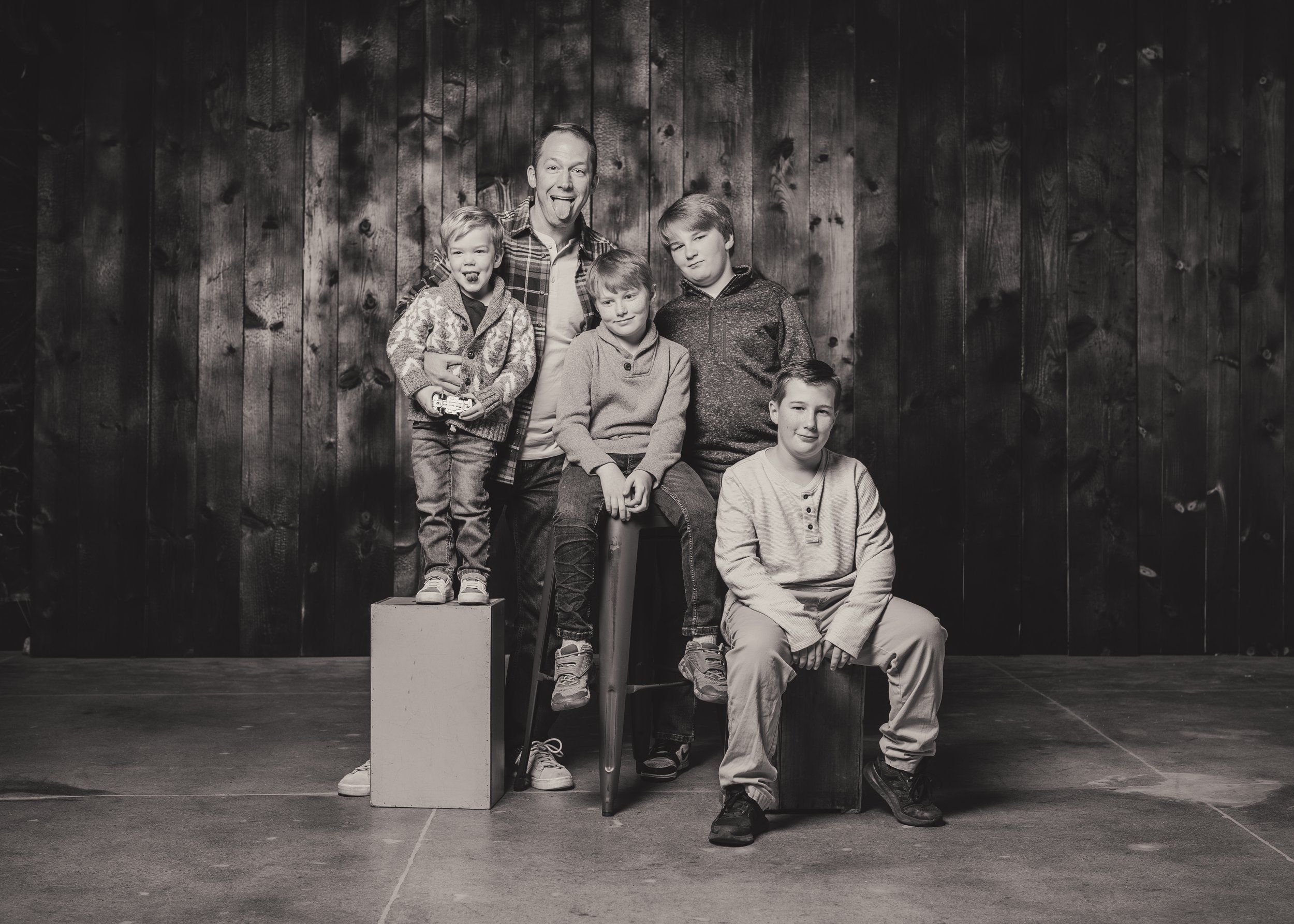 Black and white photo of a man and four boys posing together against a wooden wall. The man is standing, sticking out his tongue. One boy is sitting on a stool, another is on a box, and two are standing behind. All are casually dressed.