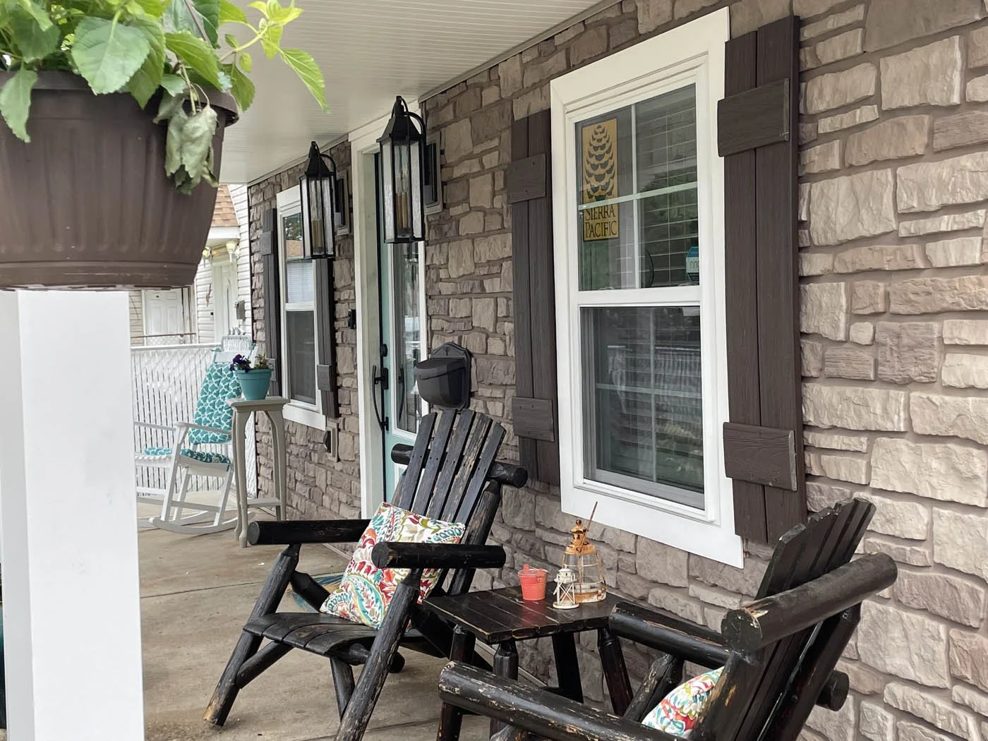 Porch with two wooden chairs, one with a colorful pillow, a small table with small lanterns and a candle, a window with brown shutters, and a stone wall. There's a hanging potted plant and a white rocking chair with a teal patterned pillow in the background.