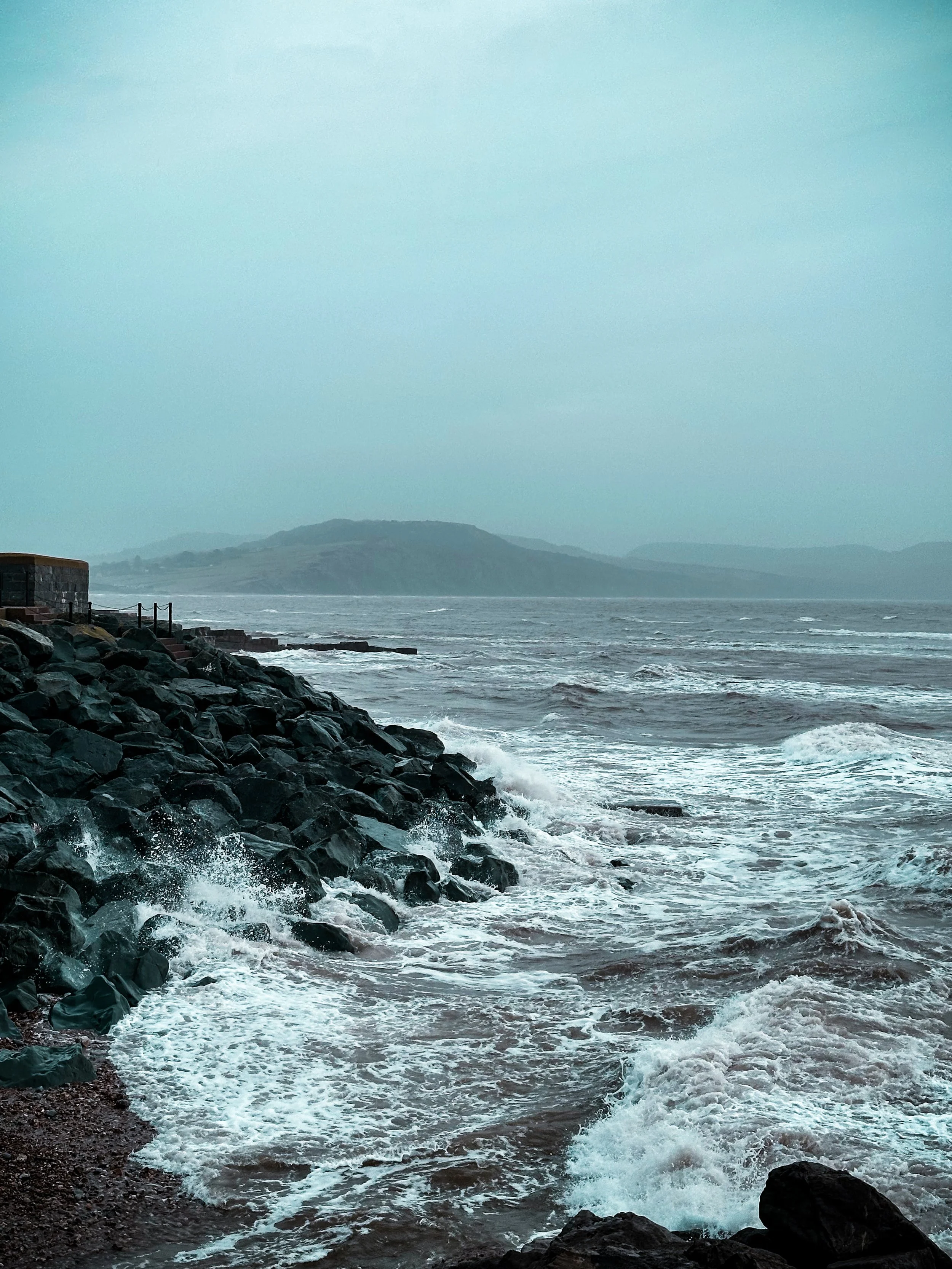 Rocky shoreline with waves crashing against the rocks, overcast sky, distant hills in the background.