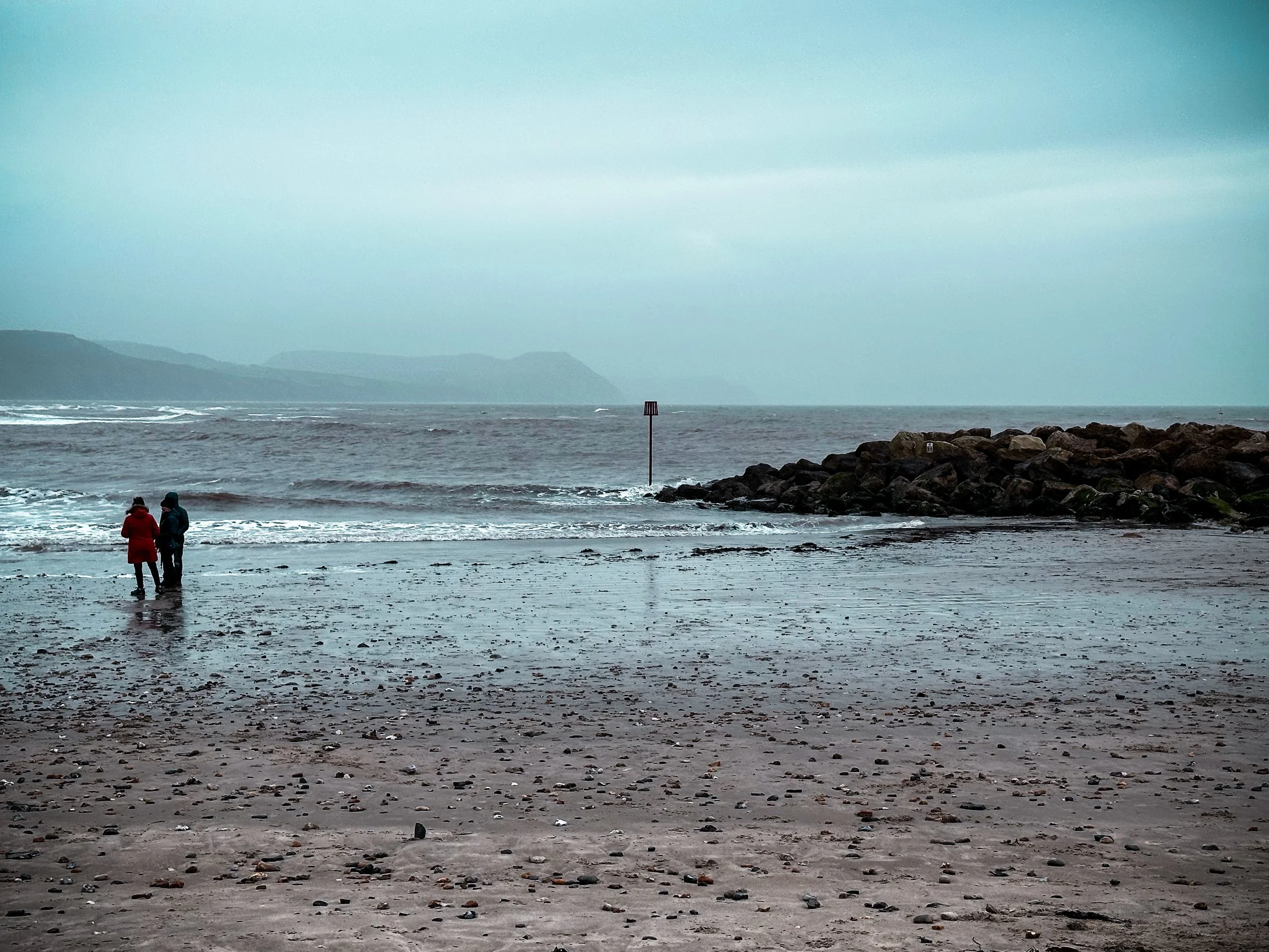 Two people standing on a beach near the water under a cloudy sky, with a rocky breakwater to the right and distant landmasses in the background.