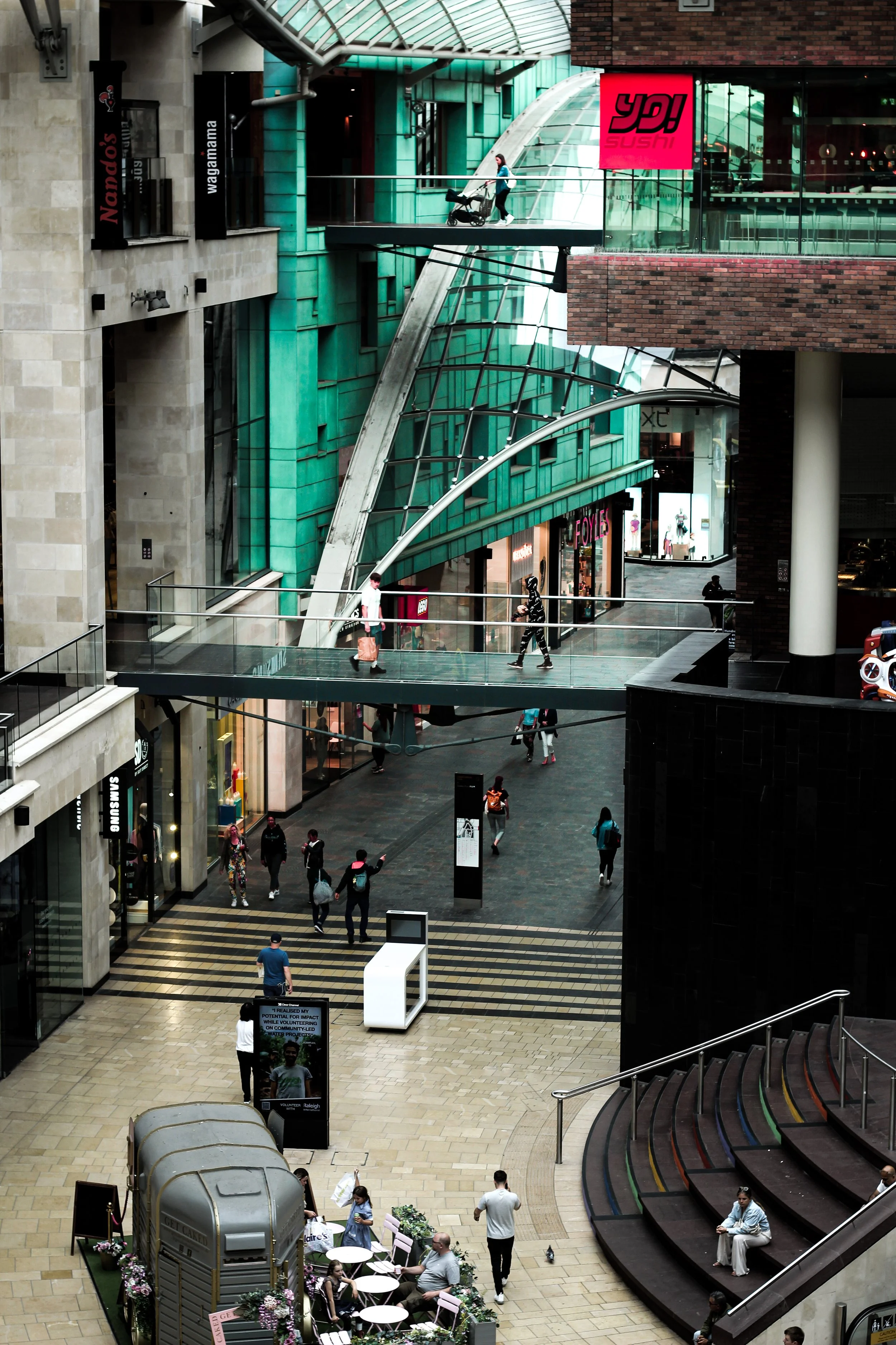 Interior of a shopping mall with multiple levels, glass walkways, and storefronts, including Nando's and Wagamama. People are walking, shopping, and sitting on stairs and benches.
