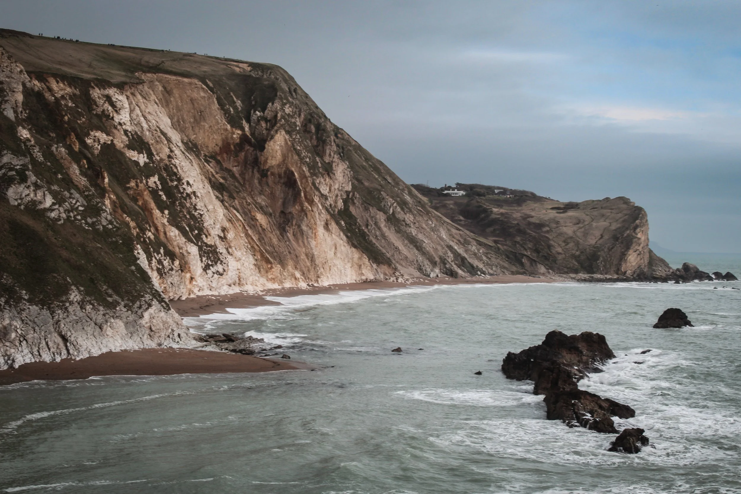 Cliffside coastline with rocky shoreline and waves crashing against rocks, overcast sky.