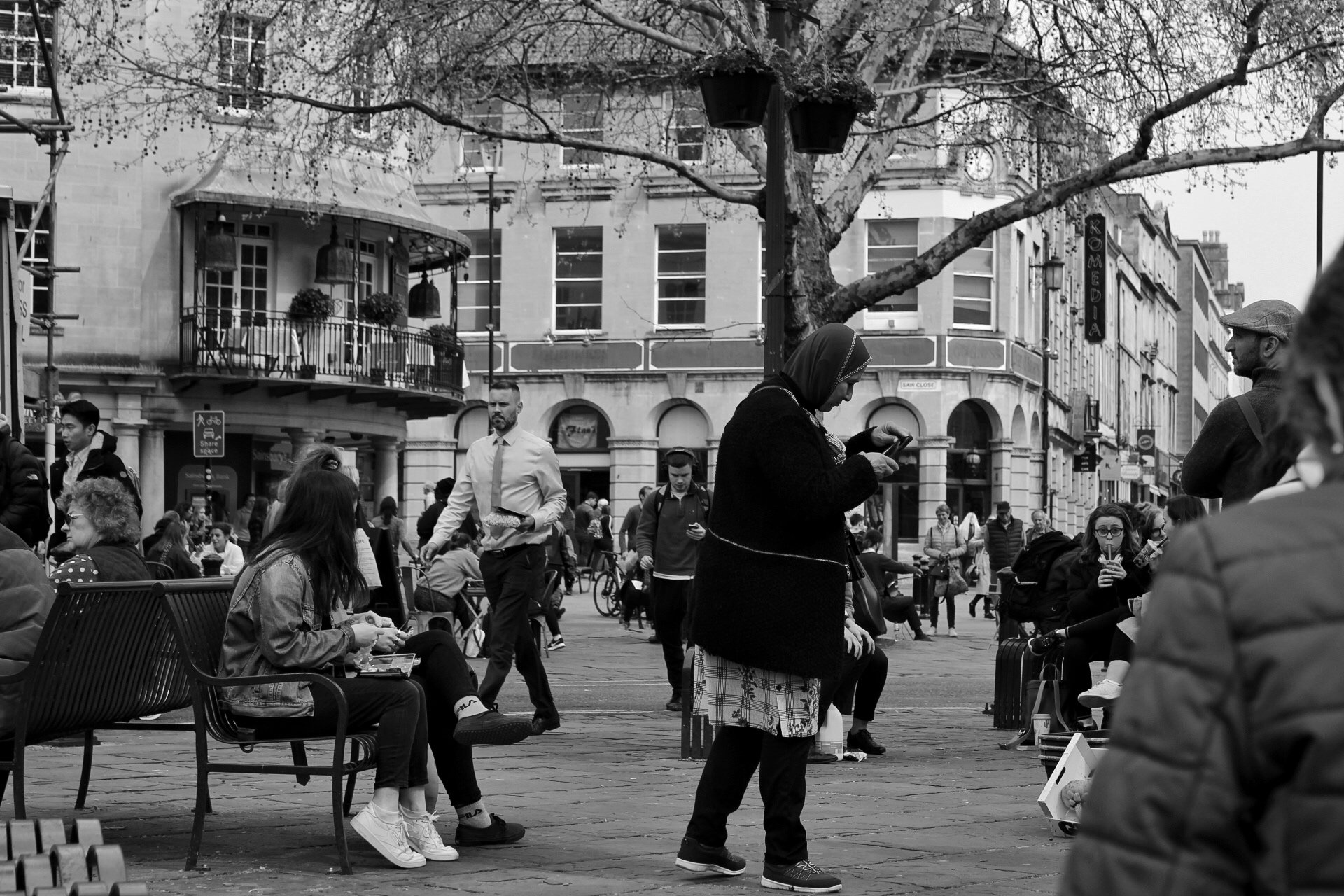 A black and white photo of a busy city square with people sitting and walking. A woman with a headscarf is looking at her phone, and other individuals are engaged in various activities, such as eating, talking, and taking pictures.