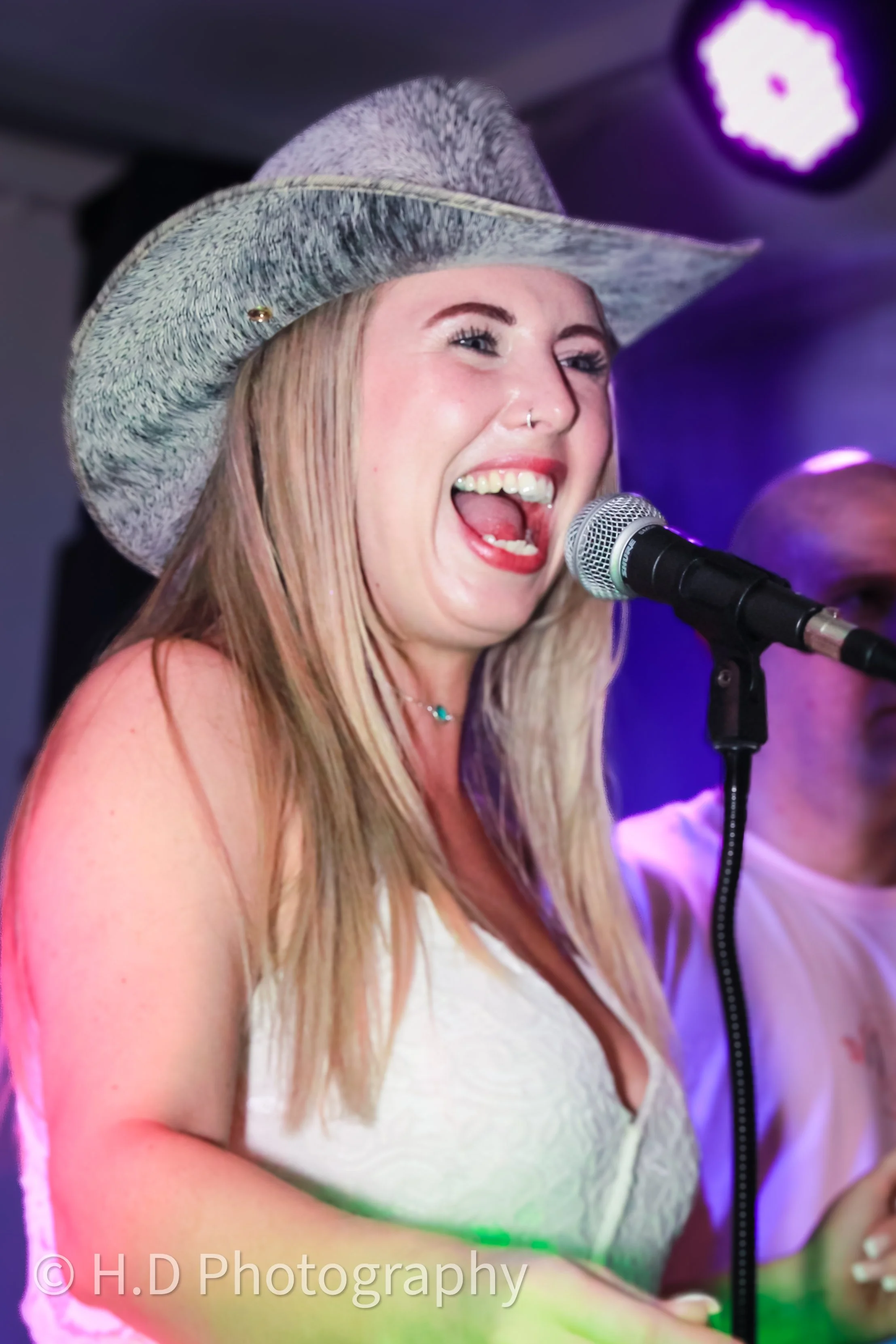A woman singing into a microphone on stage, wearing a cowboy hat, a white sleeveless top, and a necklace. She has blonde hair and is smiling.