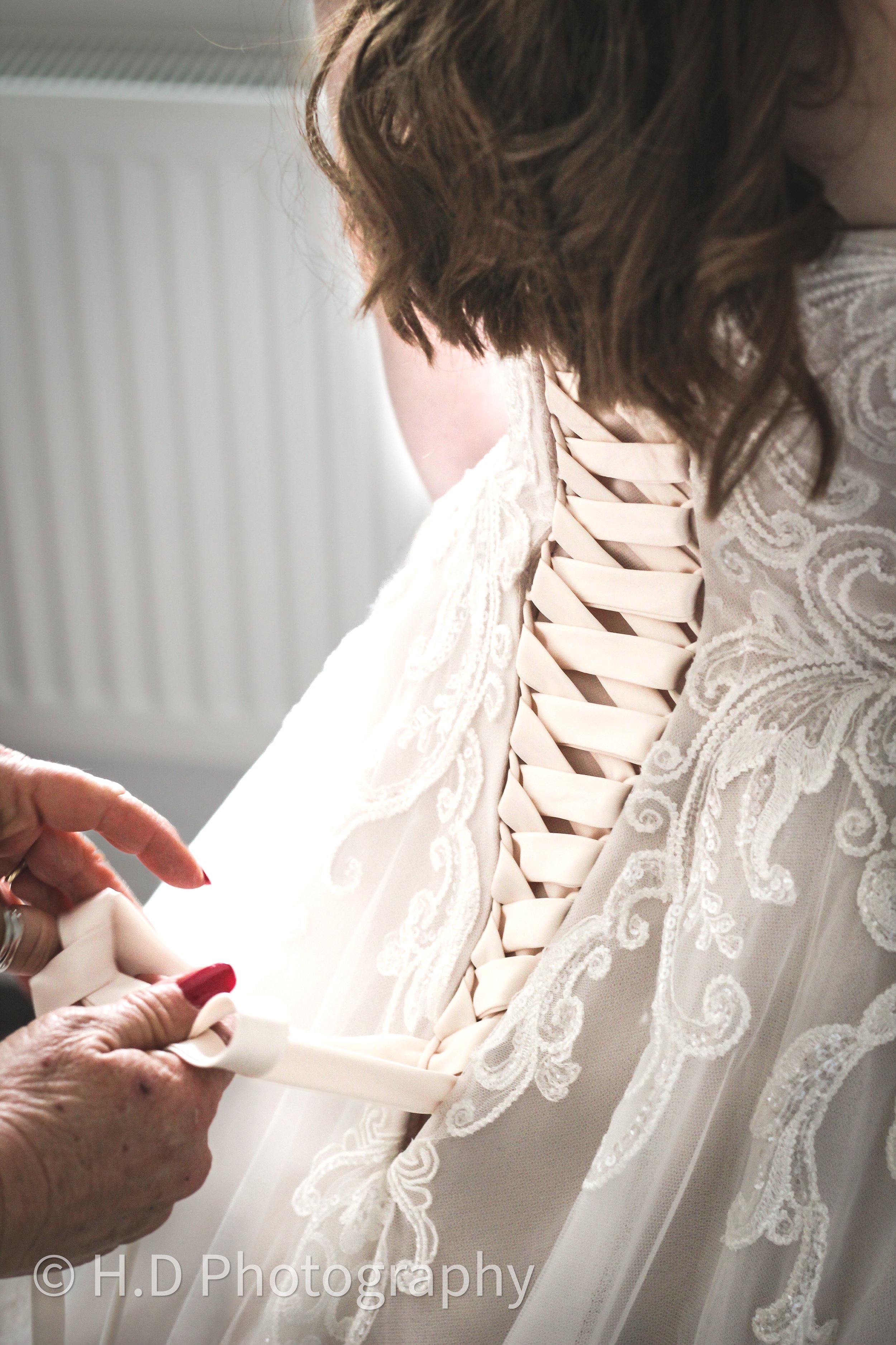 Close-up of a bride's lace wedding dress with a corset back, laced up with satin ribbon, being adjusted by someone with red nail polish, against a softly lit background.