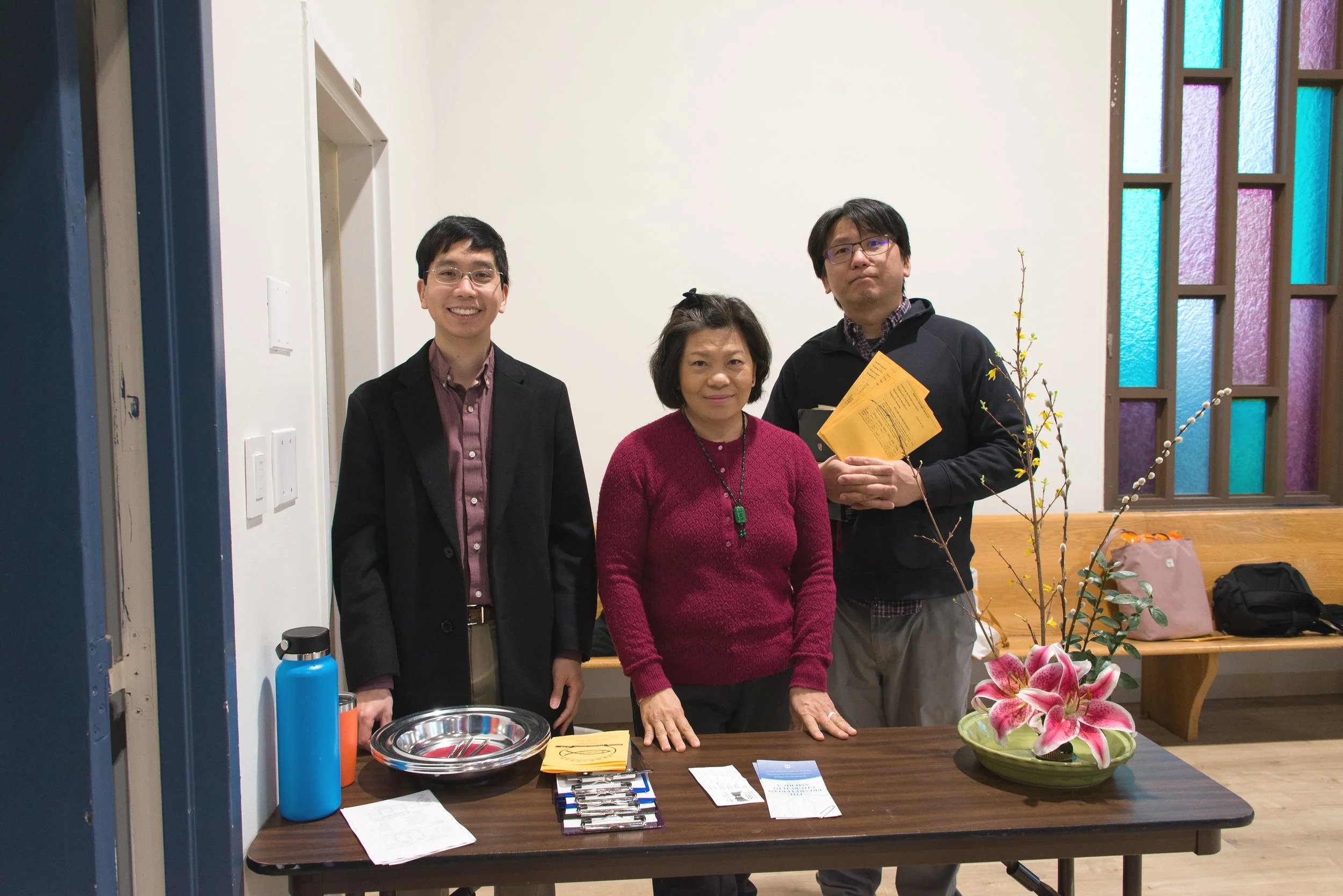 Three people standing behind a table with various items, including a flower arrangement in a green vase, papers, and a silver bowl. Two men are on either side of a woman.