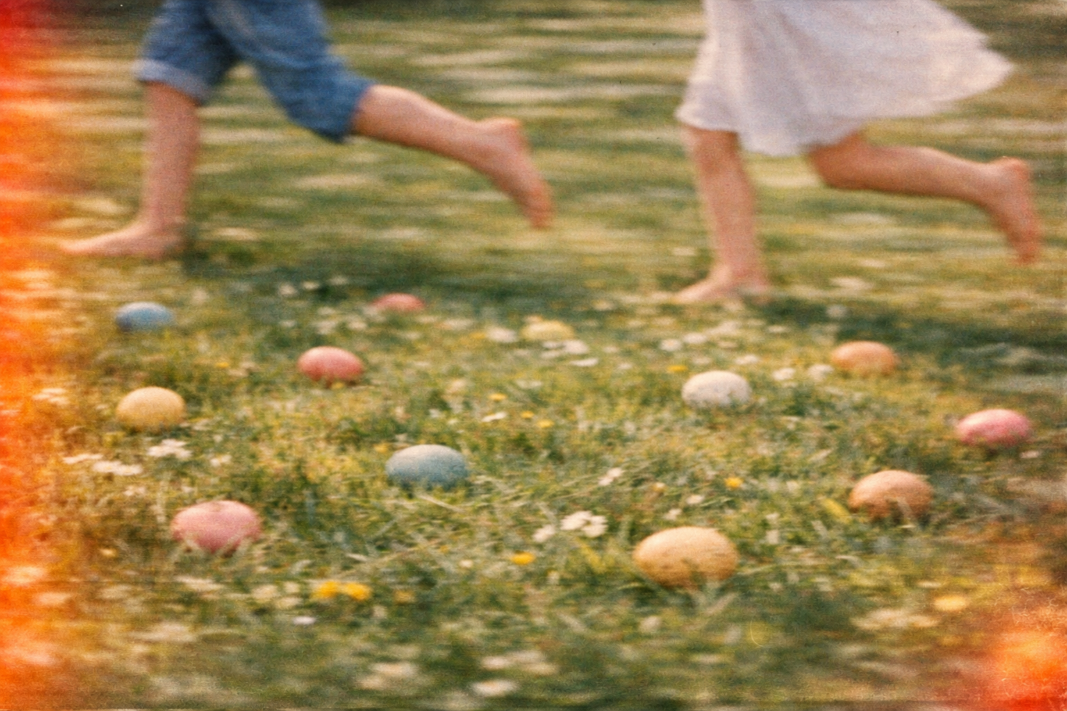 Two children barefoot running and playing outdoors on a grassy field with colorful eggs scattered on the ground, suggestive of an Easter egg hunt, during sunset or sunrise.