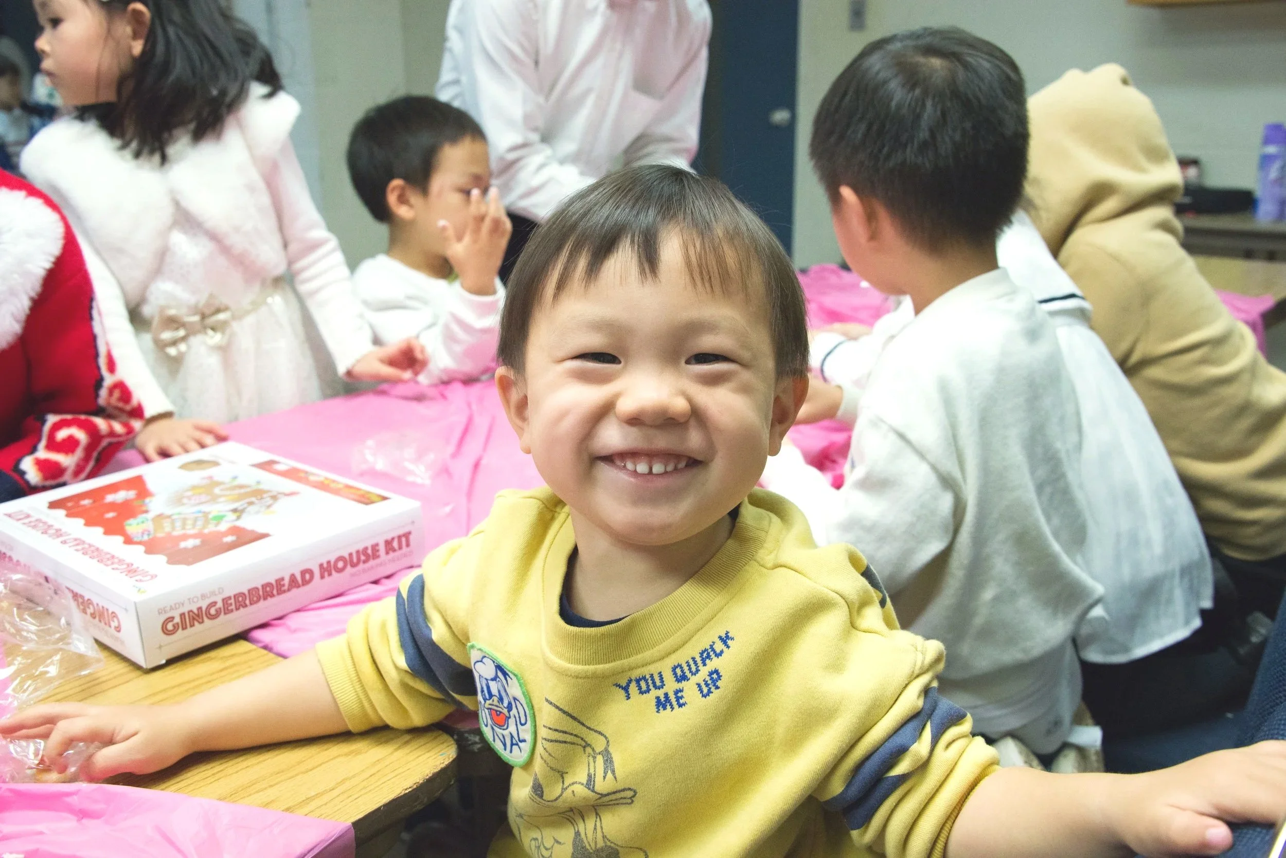 A young boy smiling at the camera during a holiday gathering, with gingerbread house kit on the table and other children in festive clothing in the background.