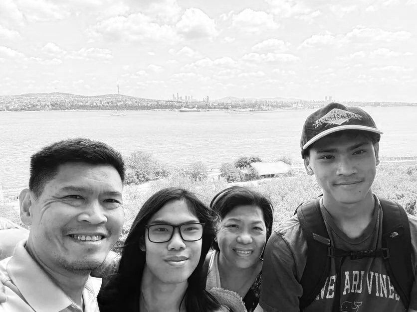A black-and-white photo of four people taking a selfie outdoors with a large body of water and city skyline in the background. The group includes two men and two women, all smiling.