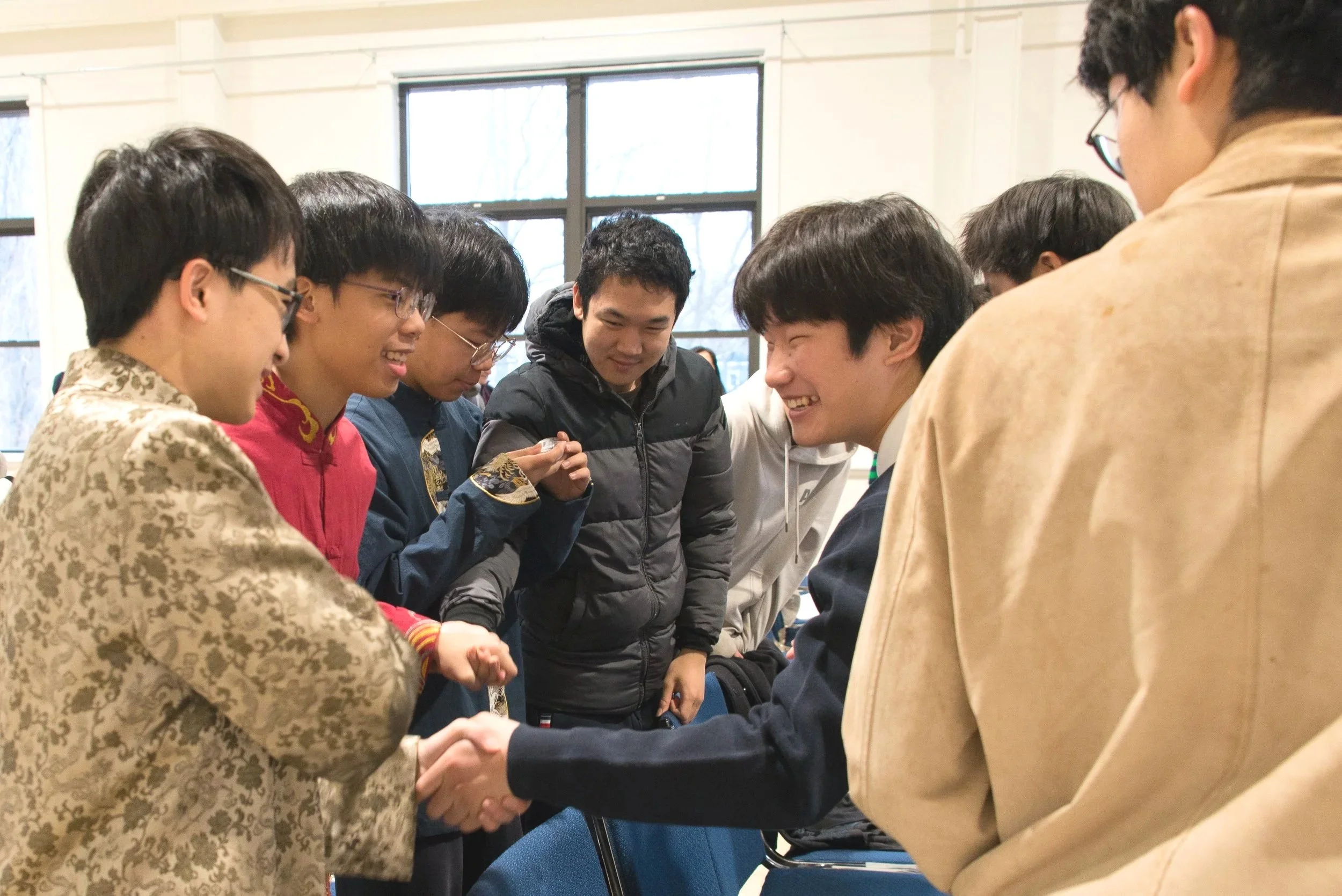 Two groups of young men shaking hands and smiling in an indoor setting with large windows.
