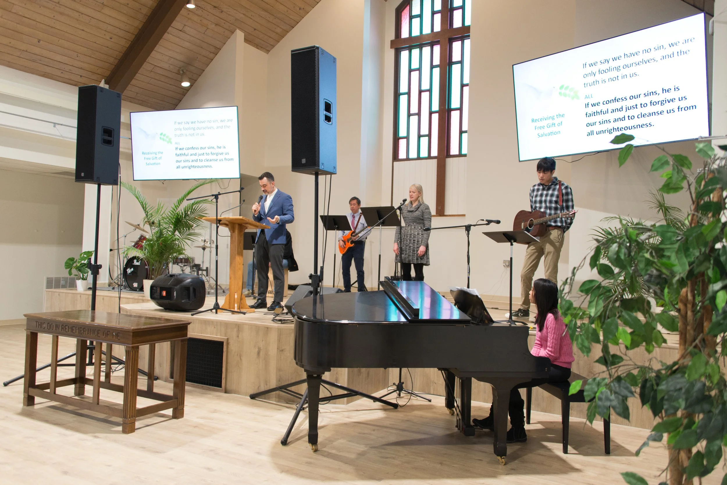 A church service with a band and a woman at the piano on stage inside a church with stained glass windows. The band includes a man in a blue suit, a woman with blonde hair, a man in a checkered shirt, and a young woman at the piano. Large screens display song lyrics.