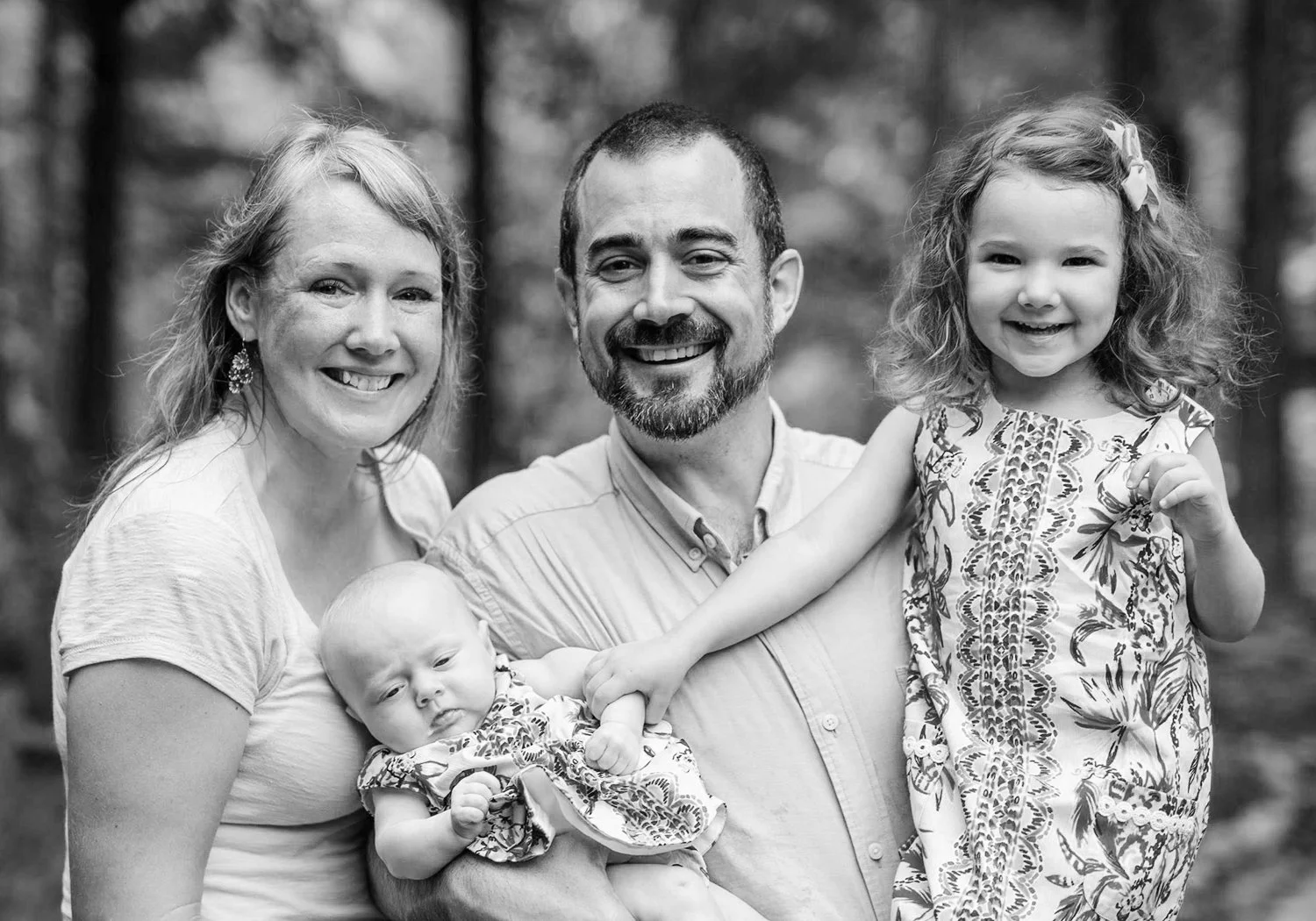 Black and white photo of a smiling family of five outdoors, including a woman with light hair, a man with a beard, a young girl with curly hair, a baby, and a woman with short hair, all smiling.