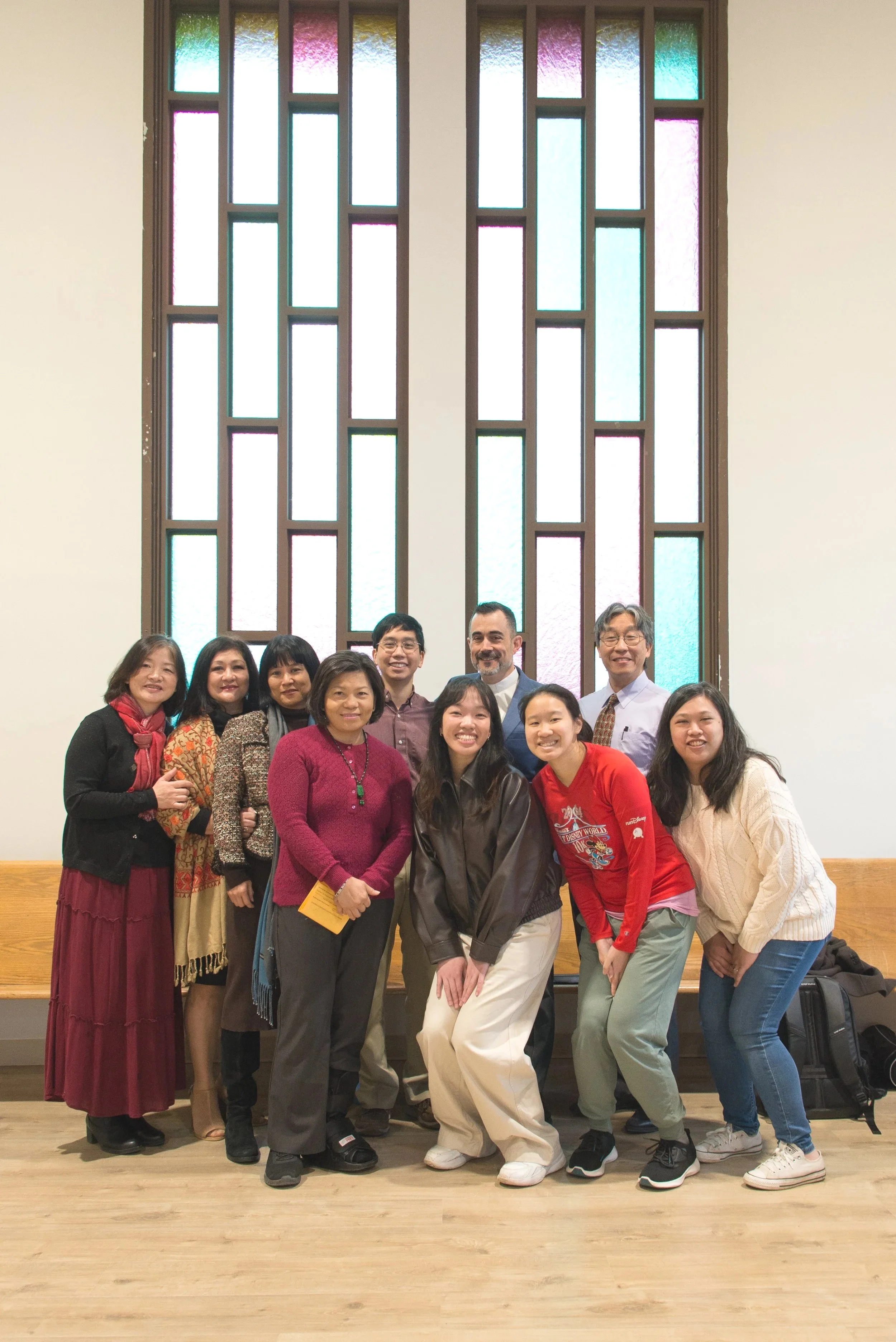Group of ten people posing in front of colorful stained glass windows inside a building.