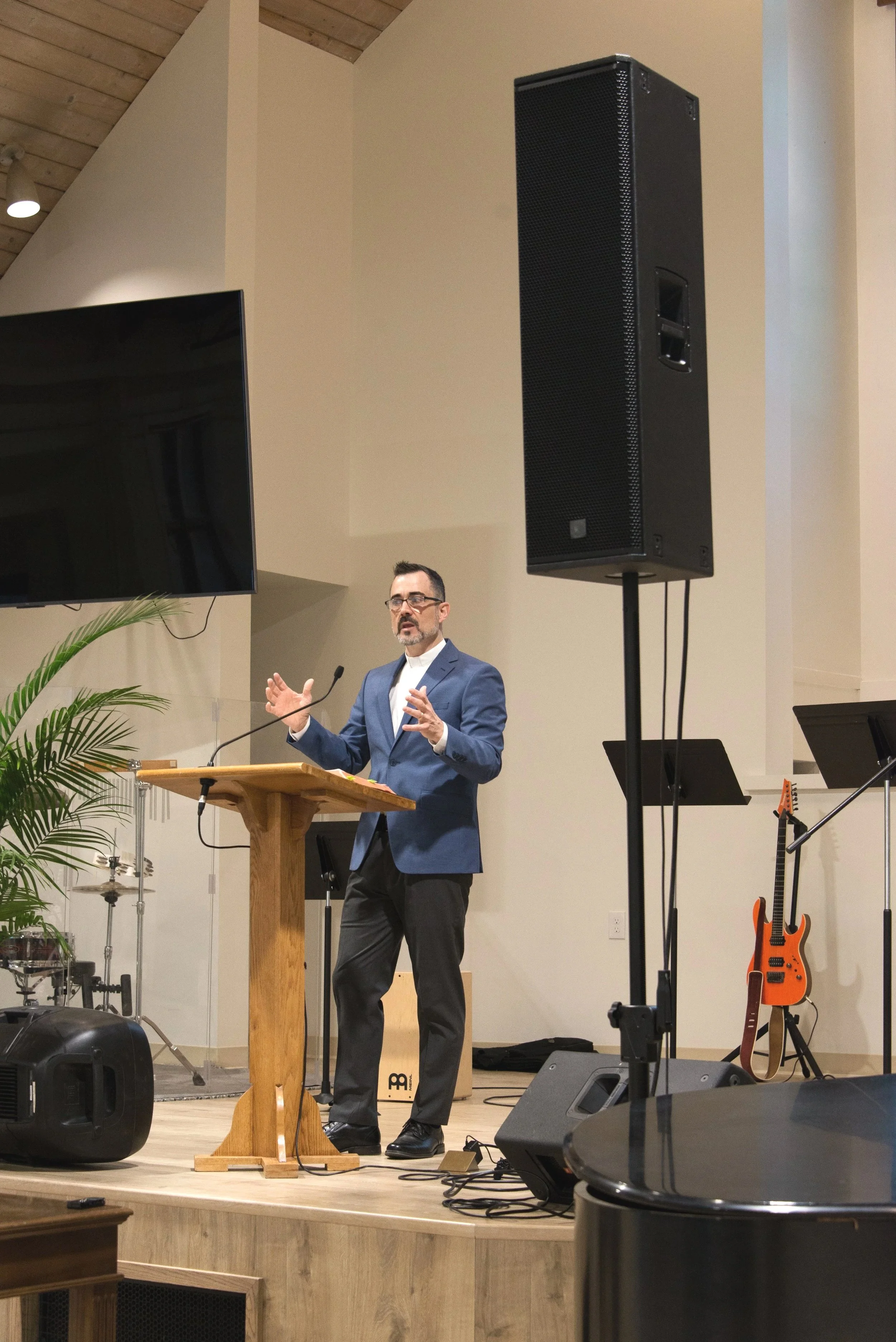 A man in a blue suit and glasses speaking at a wooden podium on a stage, with musical instruments and large speakers surrounding him.