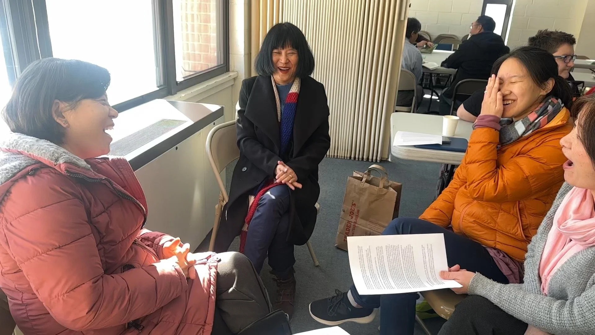 Four women sitting around a table in a bright room, laughing and talking. One woman in a red jacket is on the left, another in an orange jacket is on the right, holding a paper. The woman in a black coat and colorful scarf is in the center, smiling with her hands clasped. Two more people are in the background, working or conversing.
