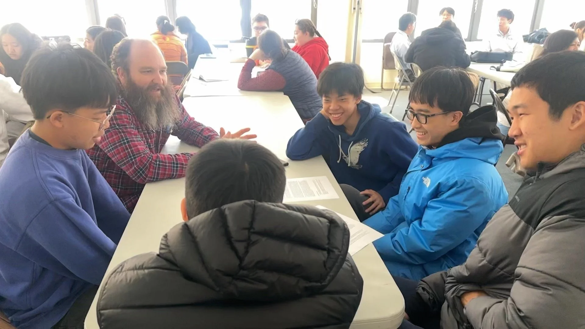 A group of students and a teacher sitting around a table in a classroom, engaging in a discussion, with some students smiling and laughing.