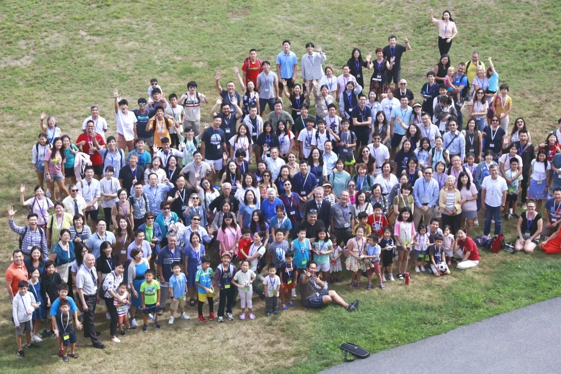 A large group of people, including men, women, and children, gathered outdoors on grass, posing for a group photo and smiling at the camera.