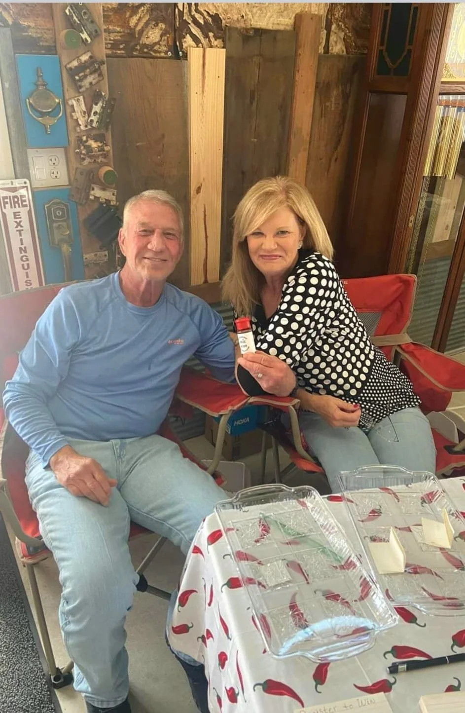 A man and woman sitting at a table decorated with a chili pepper tablecloth. The woman is holding a small bottle, possibly medicine or spice. They are smiling. The background has wooden and metallic elements, including a fire extinguisher.