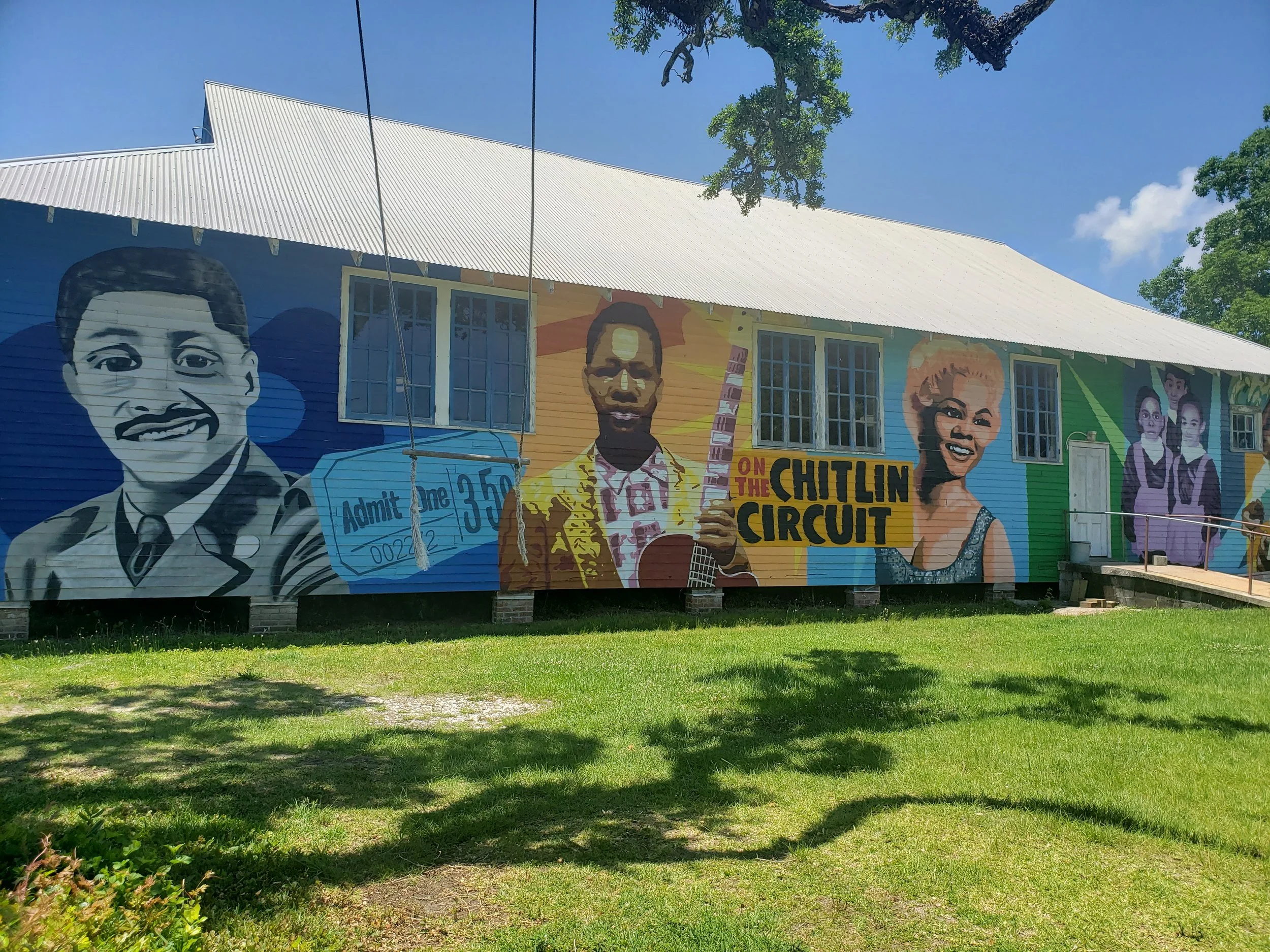 A colorful mural painted on the side of a building featuring portraits of diverse people, with a sign that says 'On the Chitlin Circuit' and a ticket with 'Admit One'.