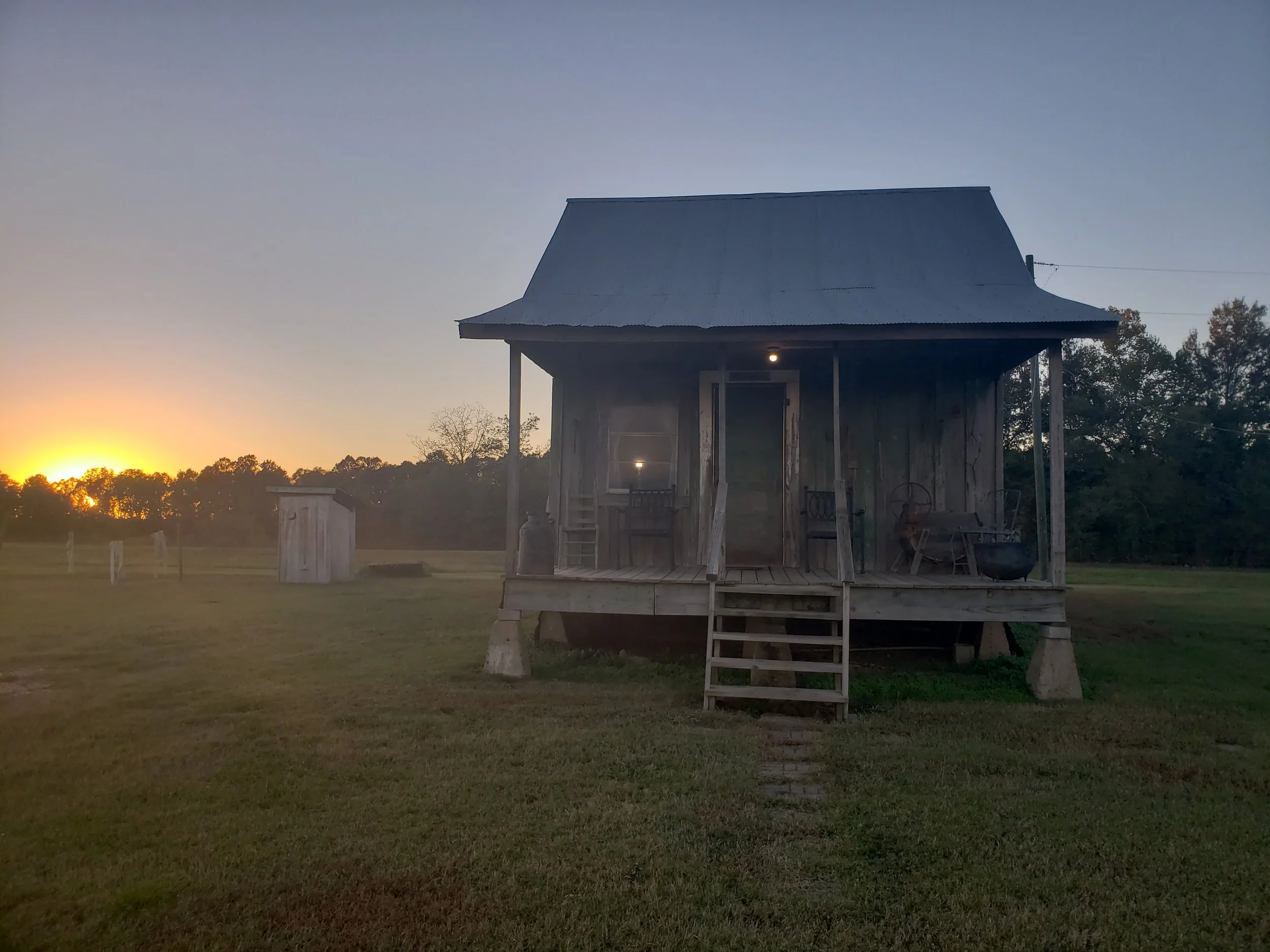 An old wooden house on stilts with a metal roof, set in a grassy field during sunset