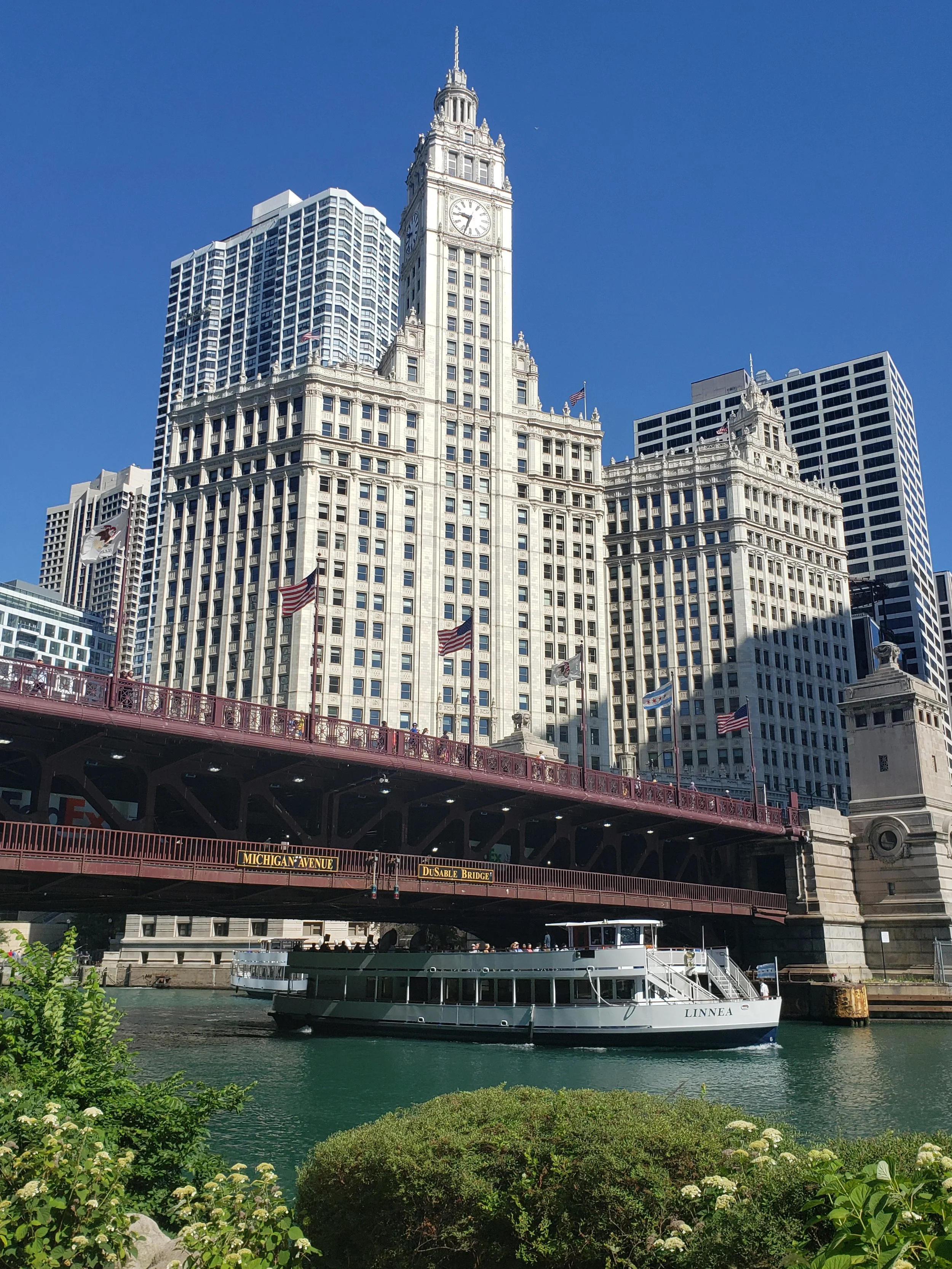 A cityscape with a historic clock tower building, modern skyscrapers behind, a bridge with American flags, a boat on the river, and greenery in the foreground.