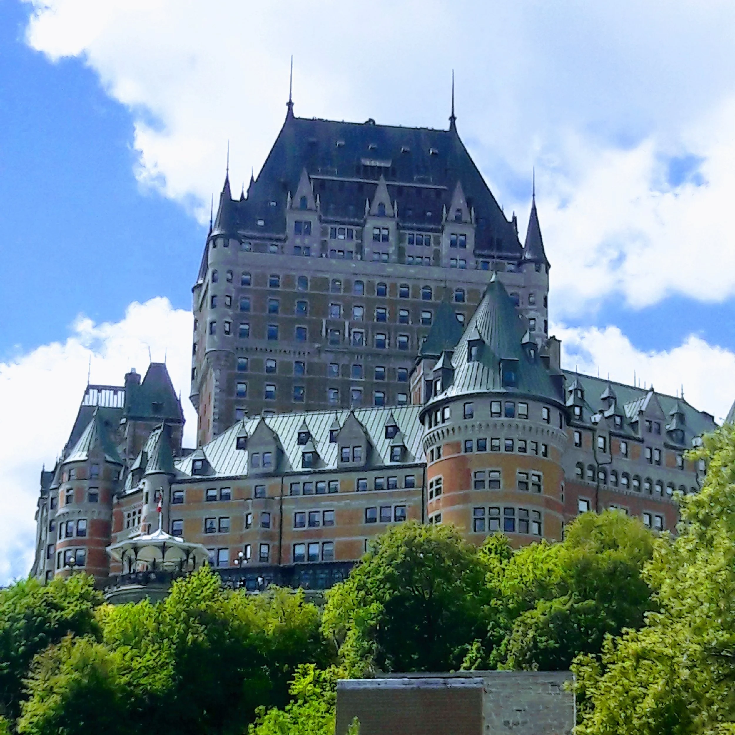 Fairmont Banff Springs Hotel in Banff National Park, Alberta, Canada, viewed from below with lush green trees in the foreground, against a partly cloudy sky.