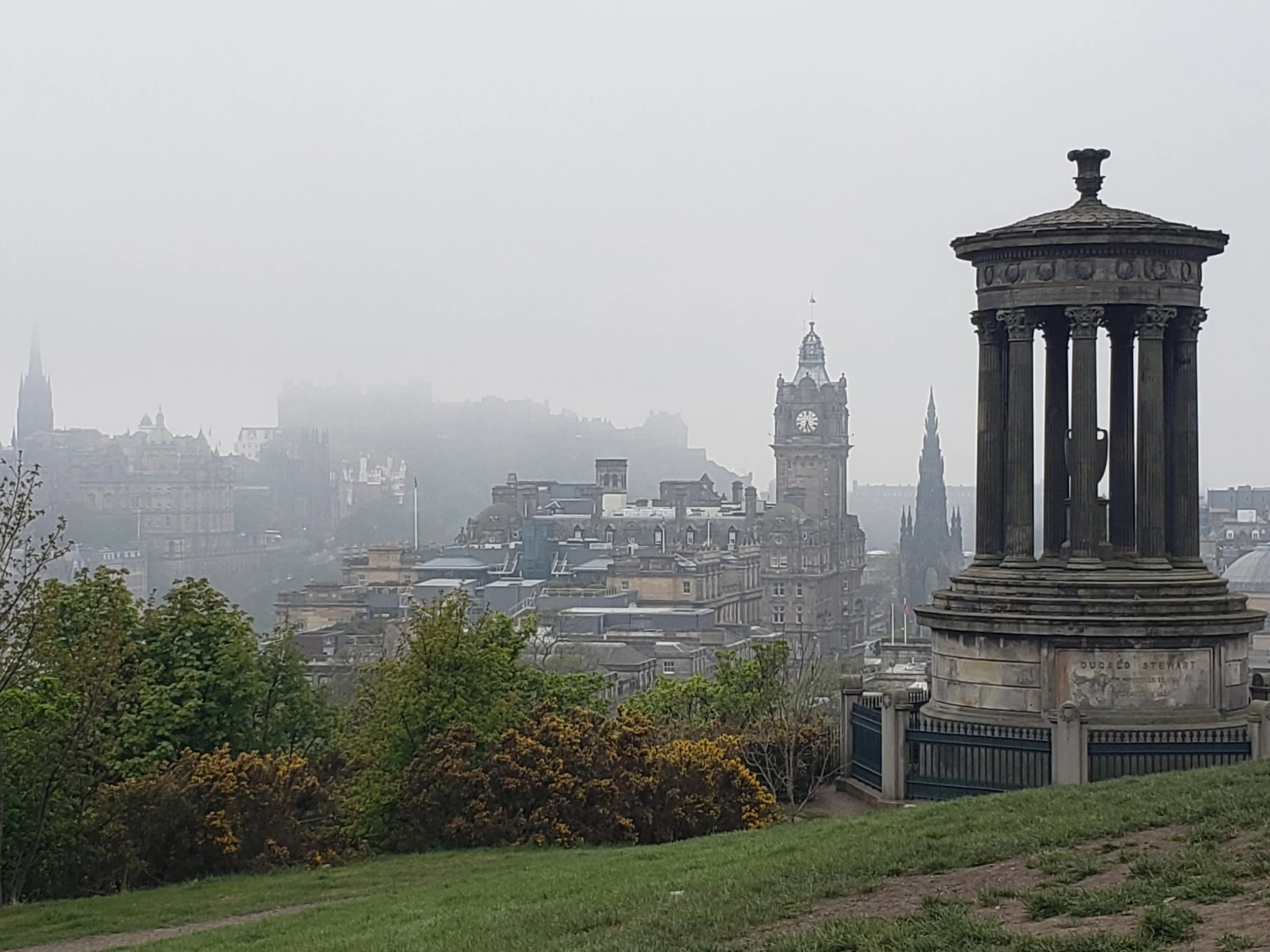 A foggy cityscape of Edinburgh with historic buildings, including the Scott Monument and Edinburgh City Hall, a green grassy foreground, and a small round monument with columns on the right side.