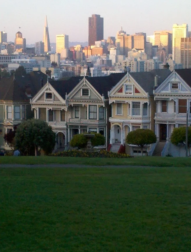 The image shows a row of Victorian-style houses with ornate details and gabled roofs, with the San Francisco skyline in the background.