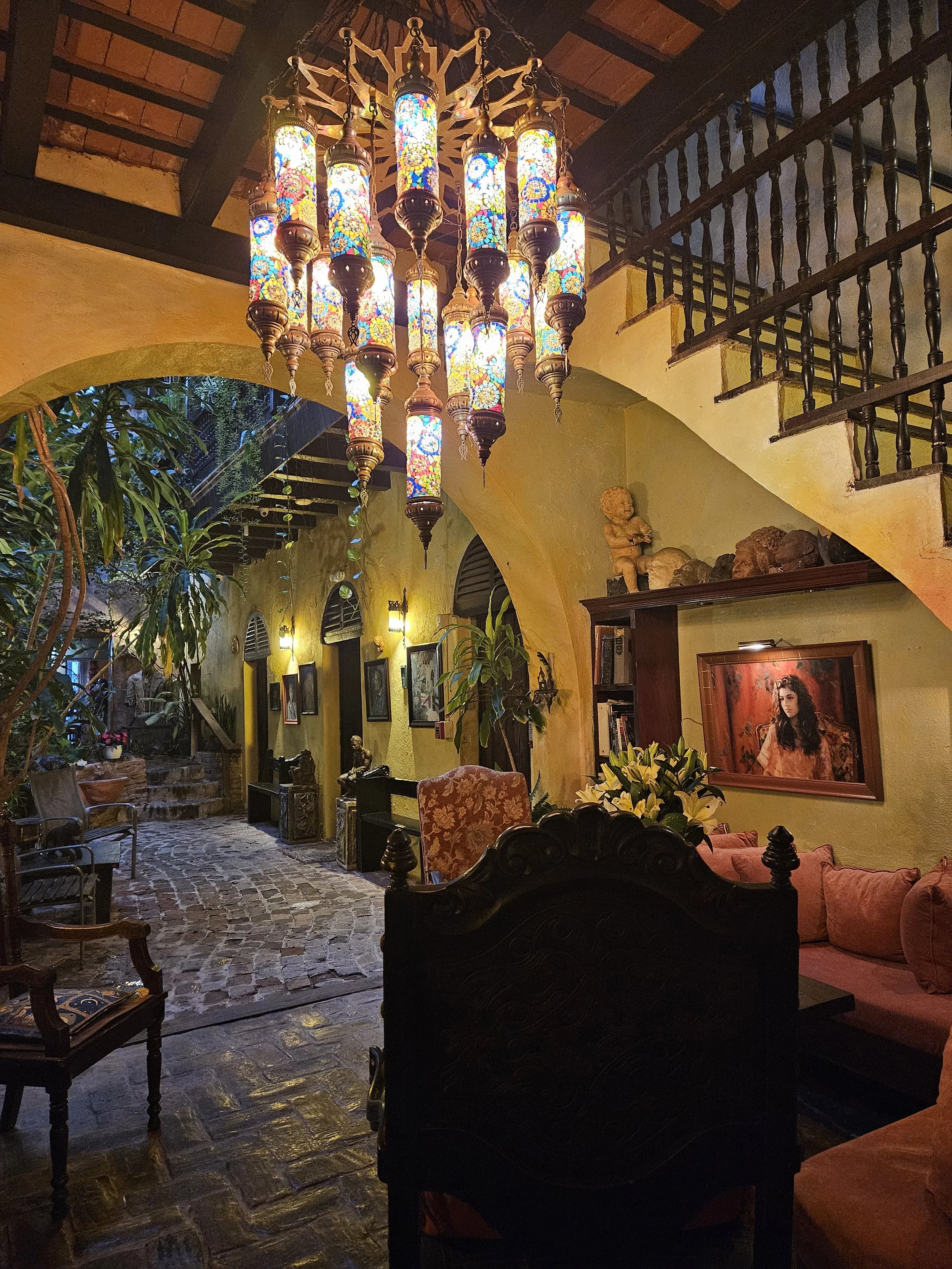 Interior of a cozy, vintage-style living room or lounge with warm yellow walls, wooden furniture, and a staircase. A large, colorful chandelier with mosaic lampshades hangs from the ceiling. There are framed pictures and decorative items on the walls