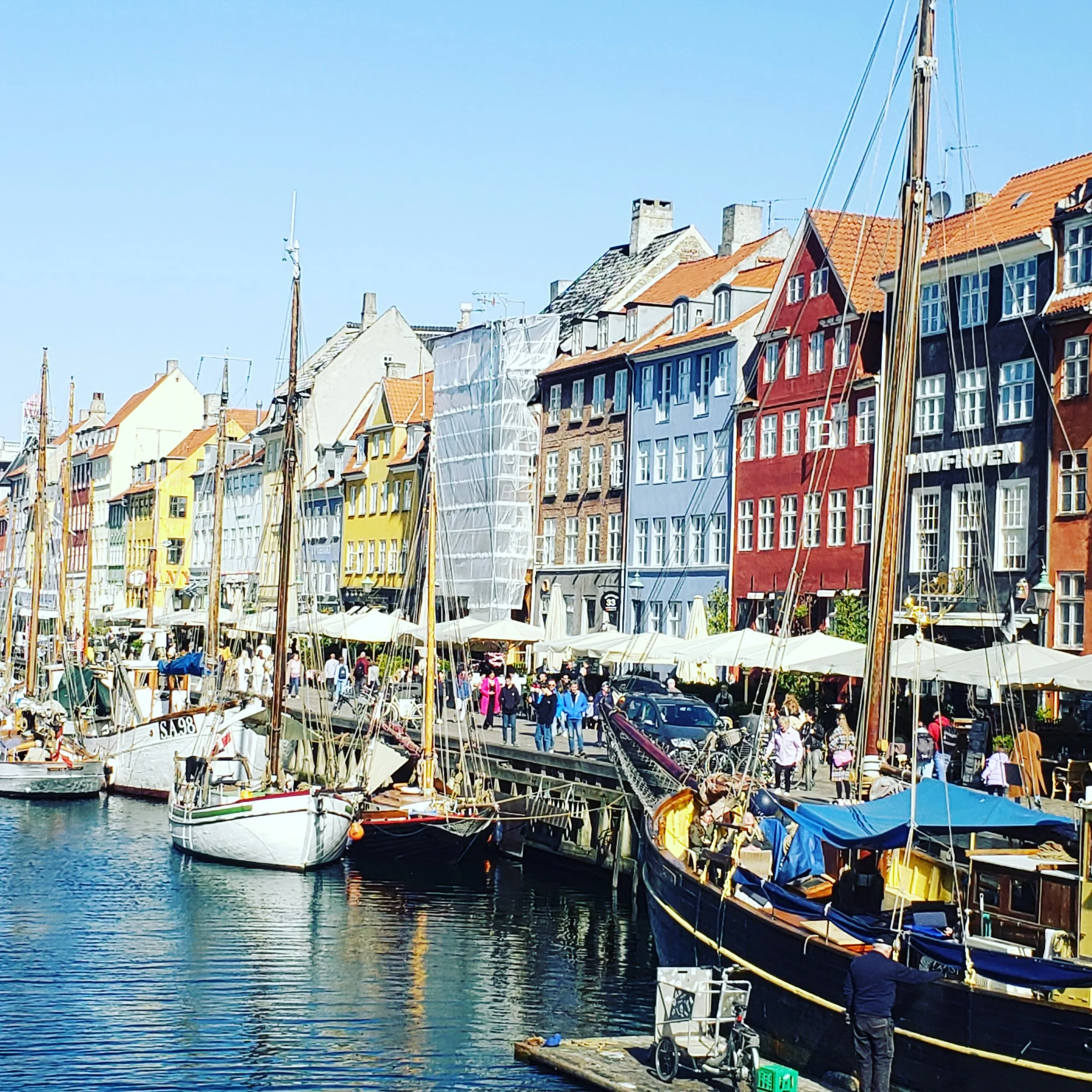 Colorful buildings along a waterfront with sailboats docked and people walking on the street.