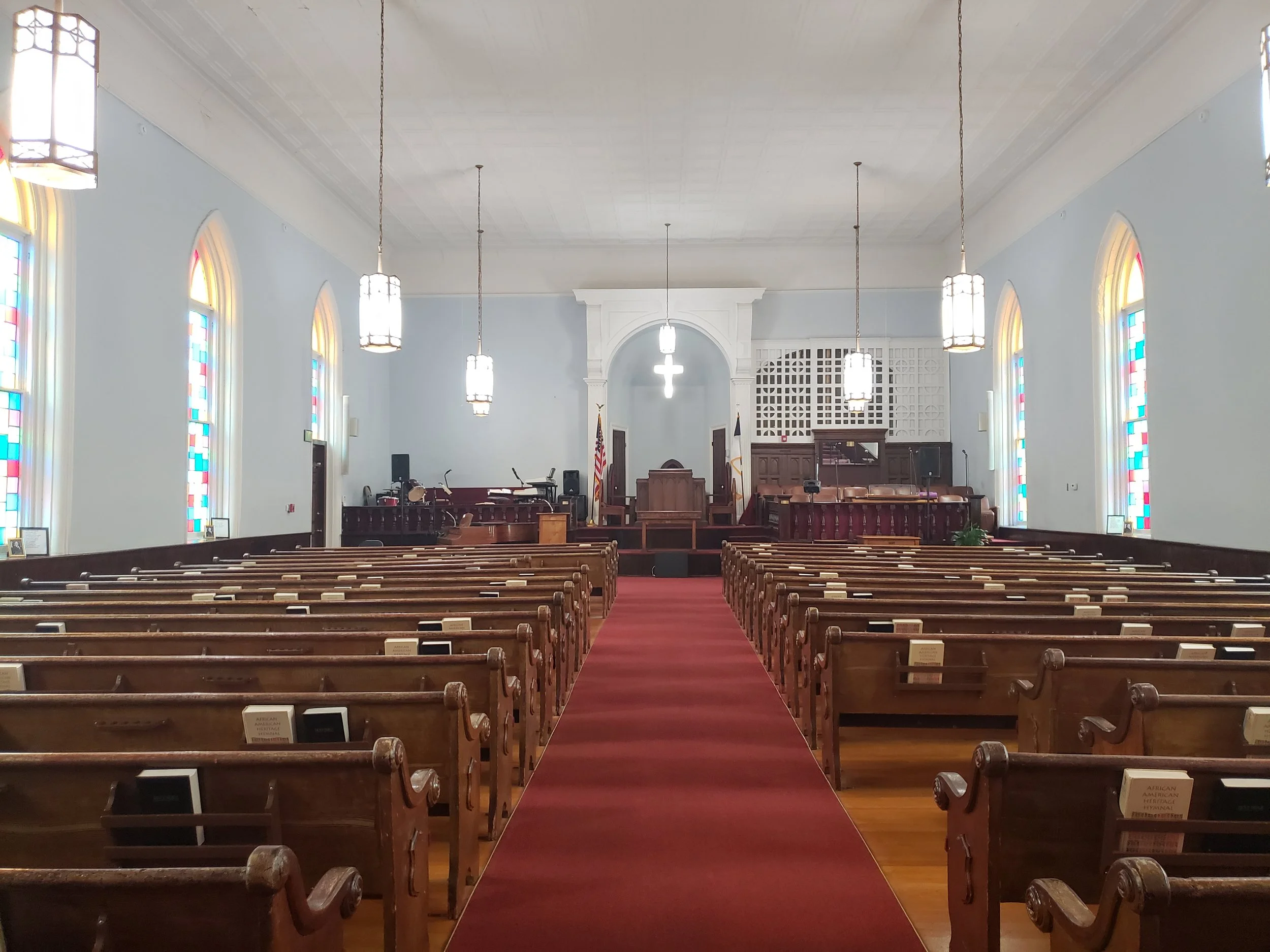 Inside a church with wooden pews, stained glass windows, and a raised choir area at the front with a cross.