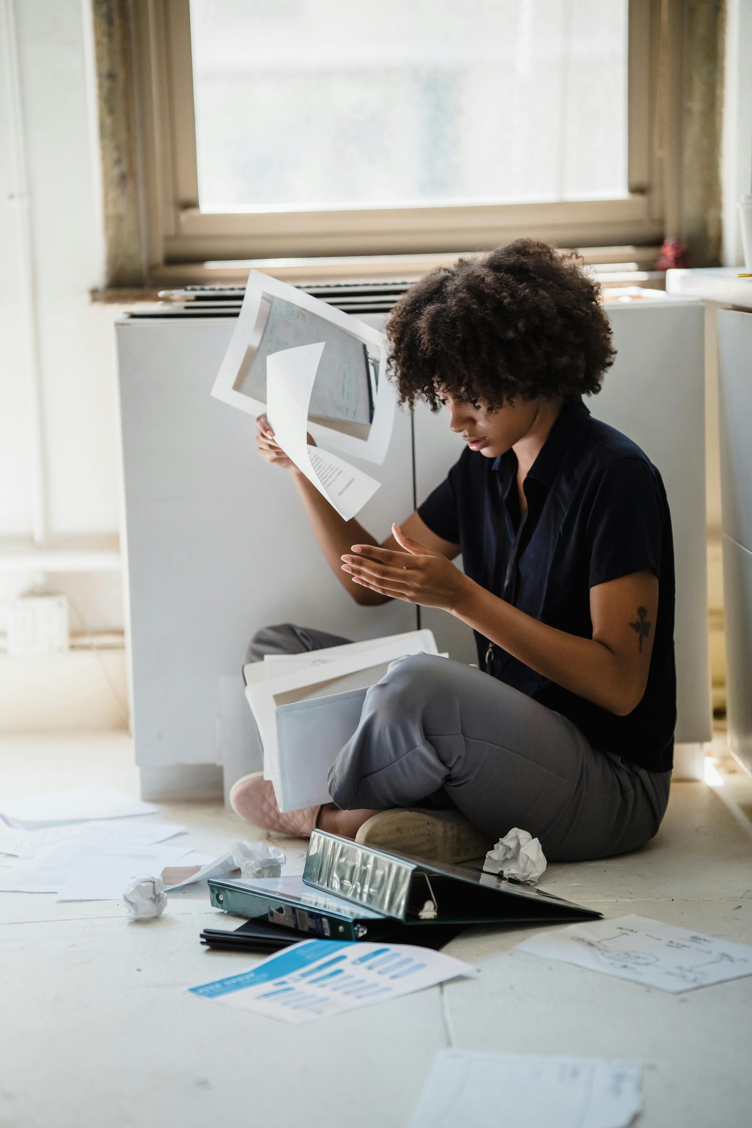 Person with curly hair sitting on the floor surrounded by crumpled papers and documents, looking at papers in their hand in a cluttered office or workspace.