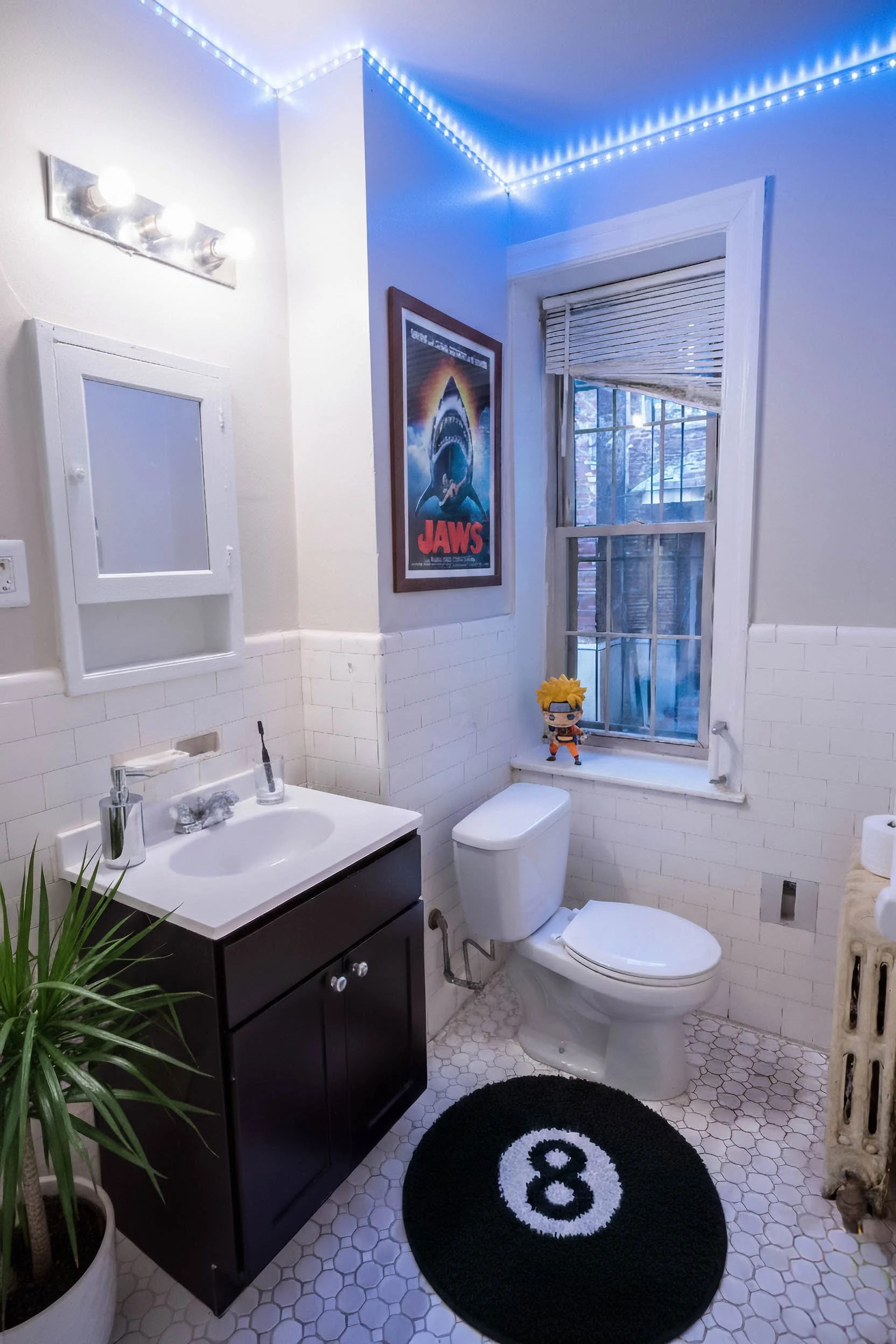 A bathroom with white tiled walls and a hexagonal tile floor. It has a black vanity with a white sink, a mirror with three lights above, a toilet, a window with blinds, a small potted plant, a framed "Jaws" poster, a figurine on the windowsill, and a