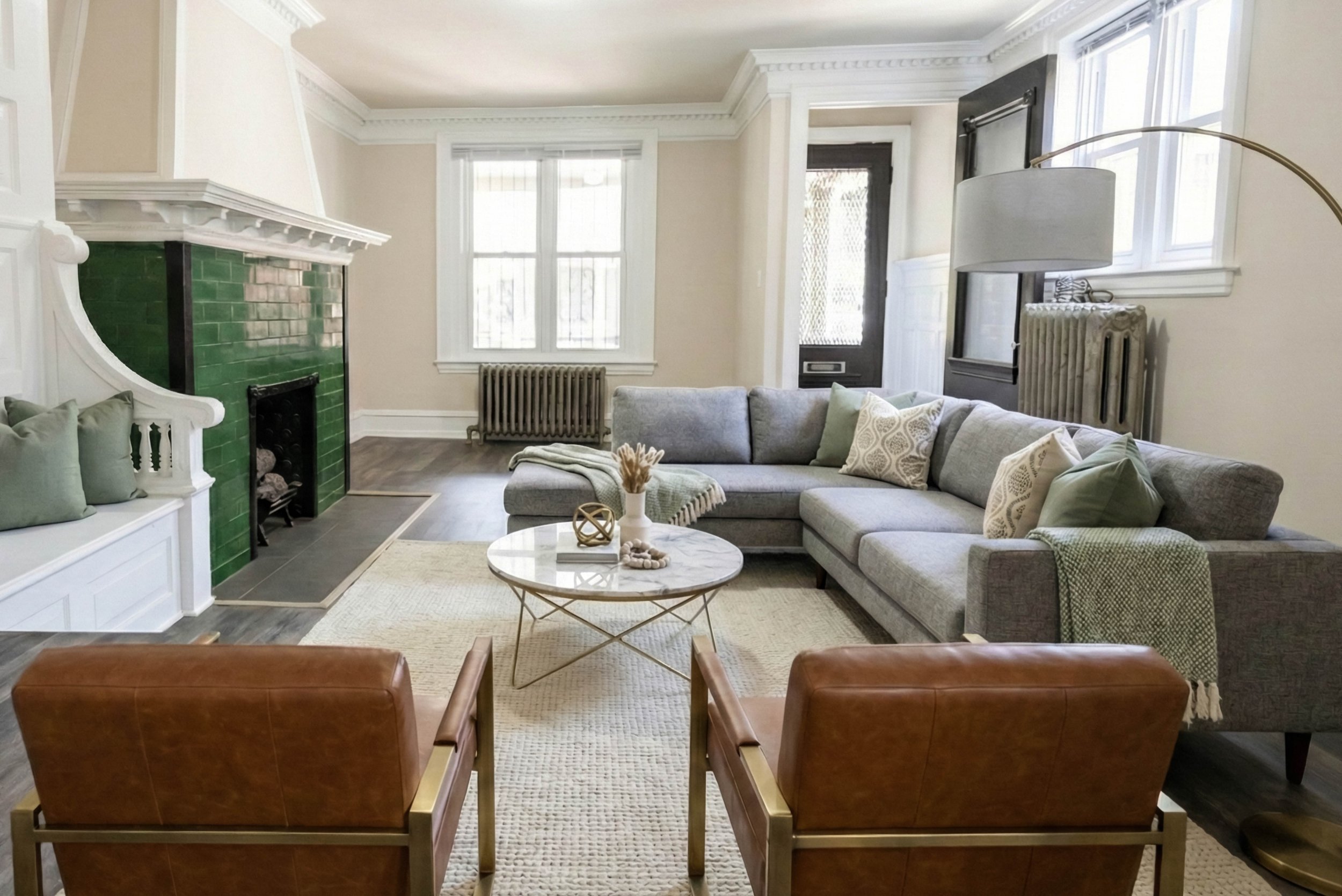 Living room with a gray sectional sofa, two tan leather chairs, a marble coffee table, a fireplace with green tiles, and large windows.