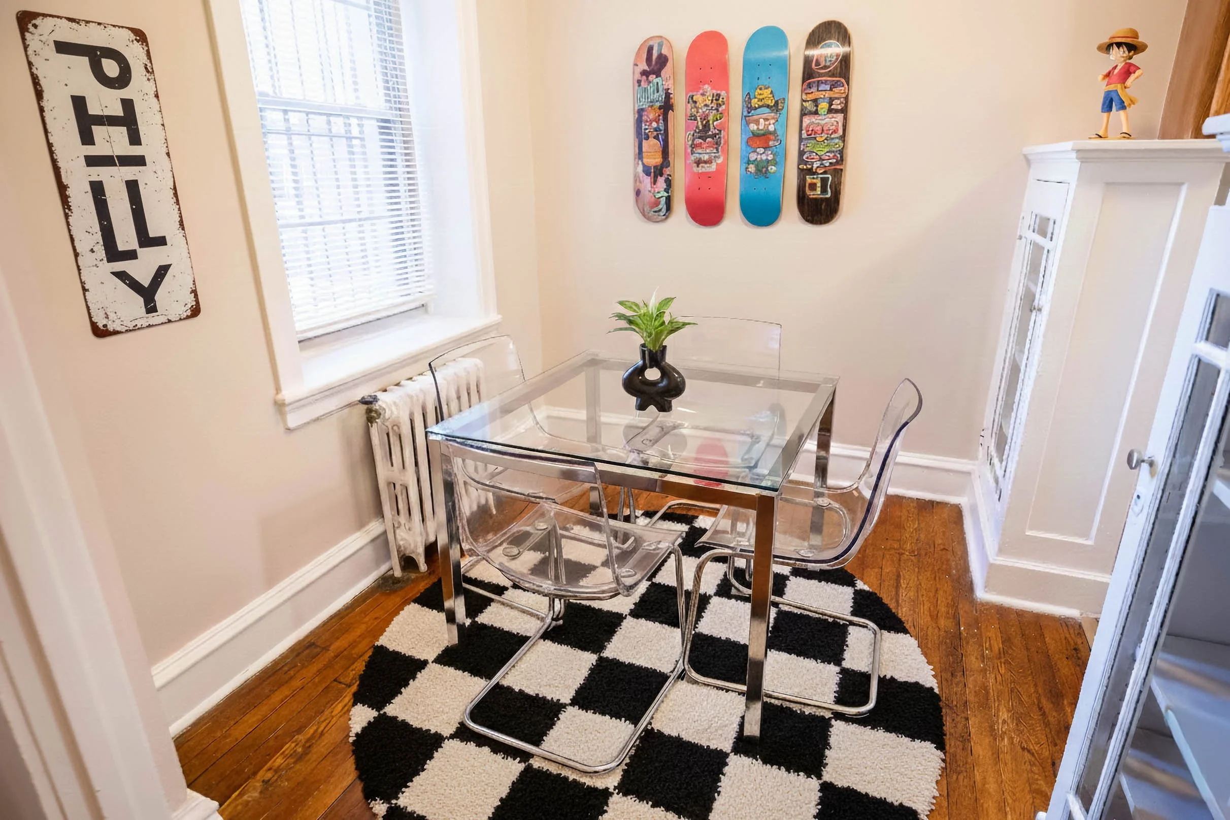 A dining room with a glass-top table, three clear acrylic chairs, a black and white checkered rug, a window with white blinds, skateboards on the wall, and a white cabinet with a figure on top.